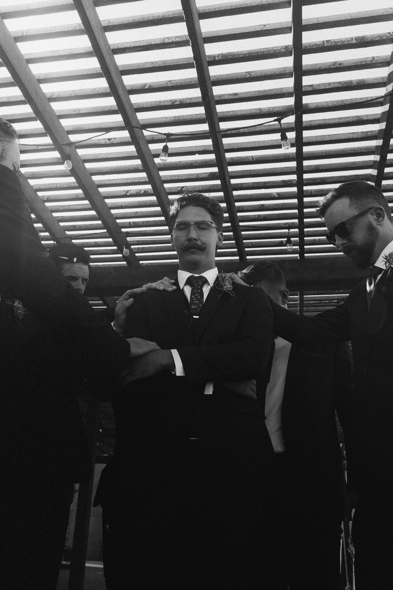 Black and white photo of a man in a suit with crossed arms surrounded by others under a striped ceiling.