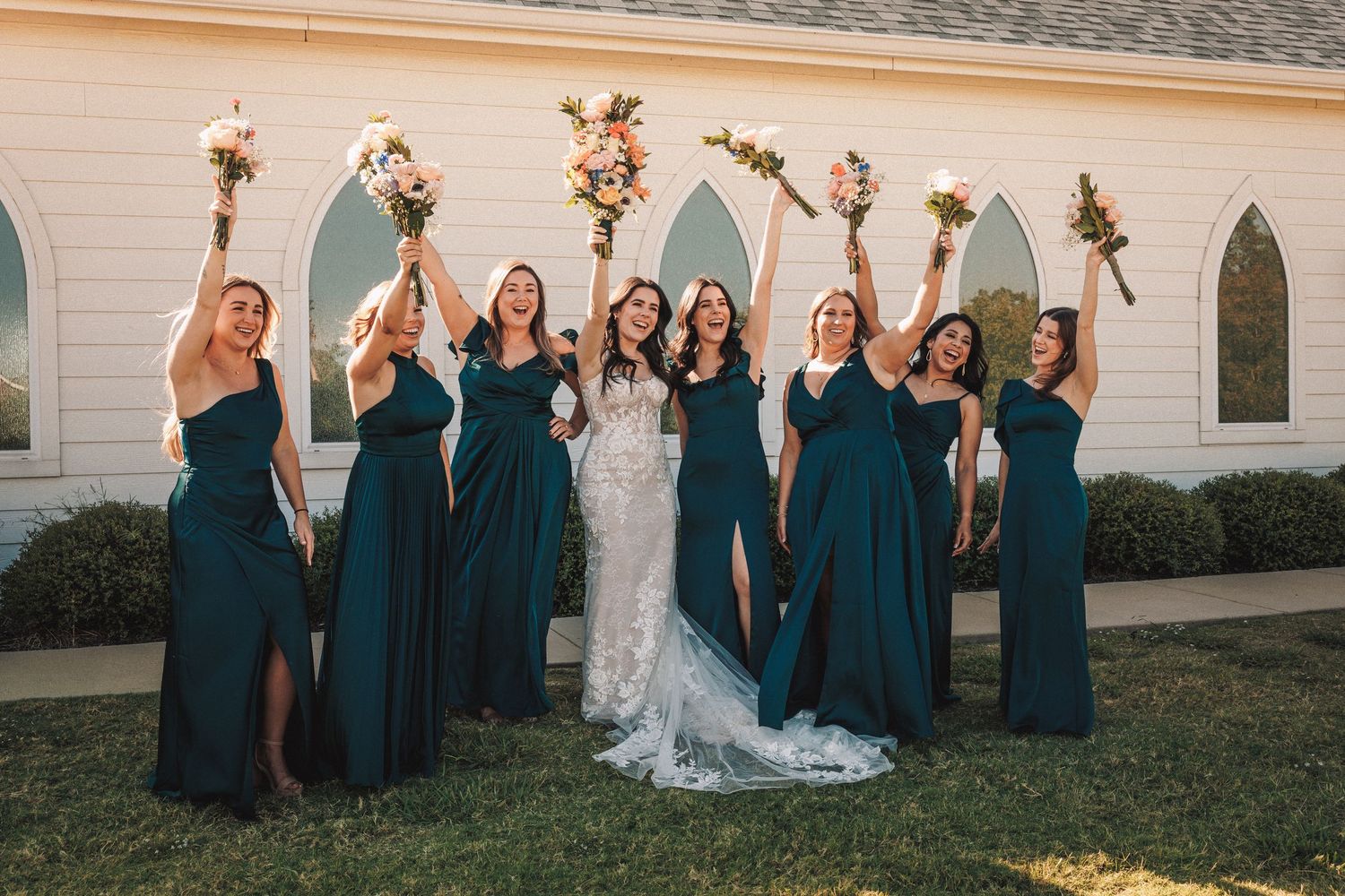Bride in lace gown with seven bridesmaids in emerald dresses raising bouquets outside a white chapel.