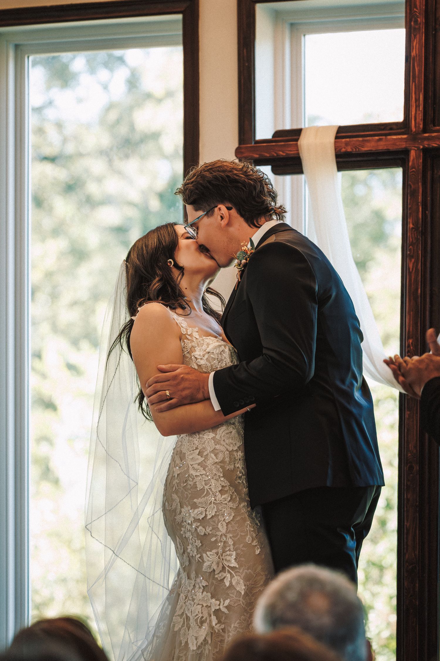 Bride and groom share their first kiss beside a wooden cross with sheer white draping at the altar.
