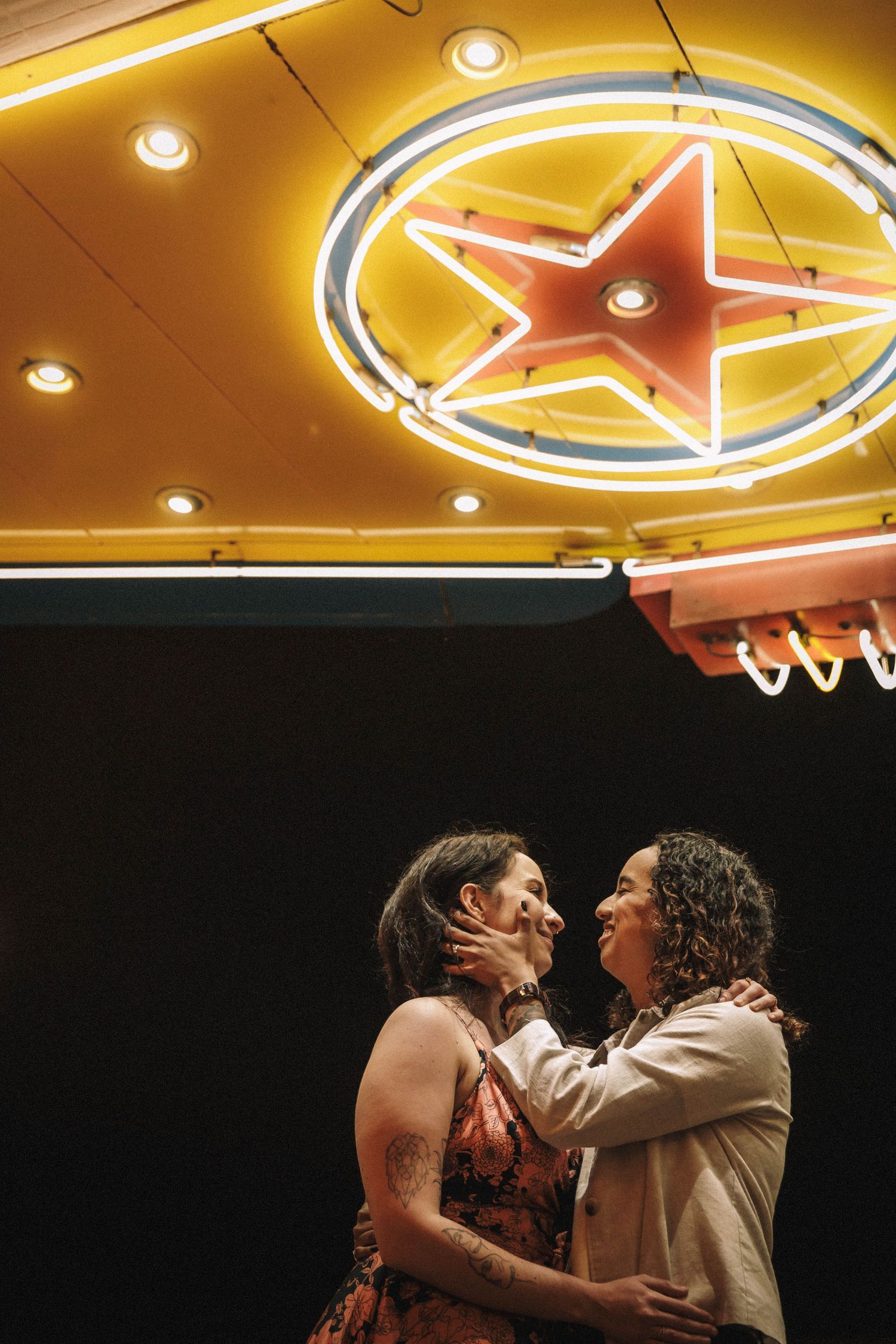 Two women share a romantic moment under a glowing neon star sign at a vintage theater.