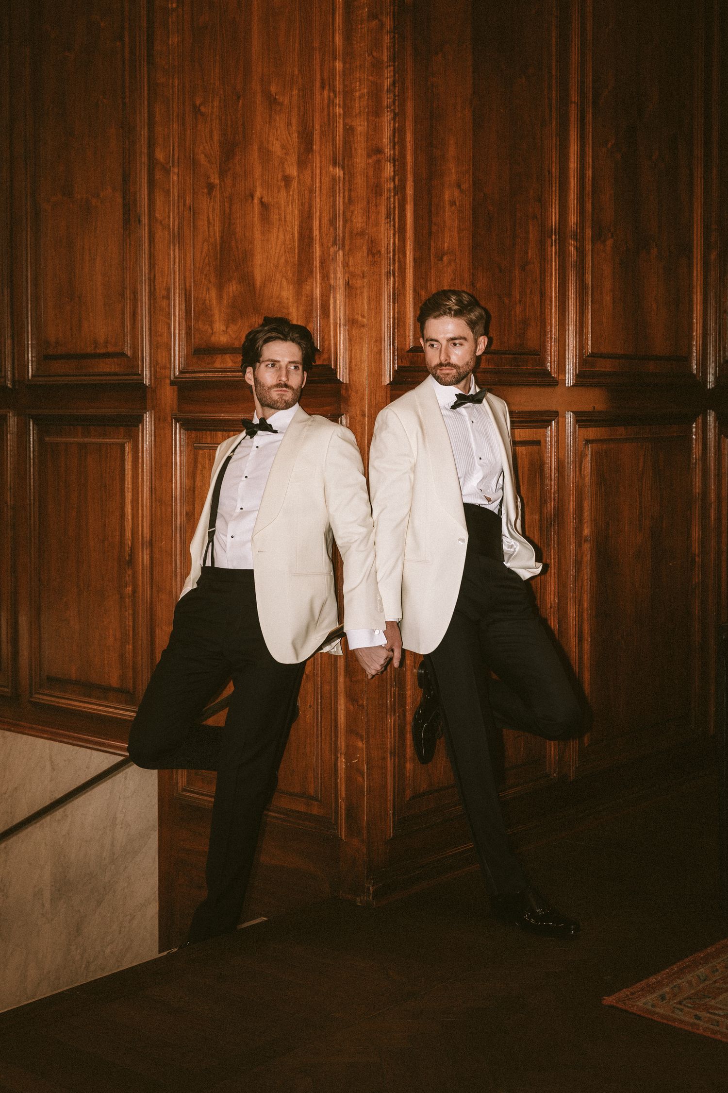 Two grooms in white tuxedo jackets pose back-to-back against elegant wood-paneled walls.