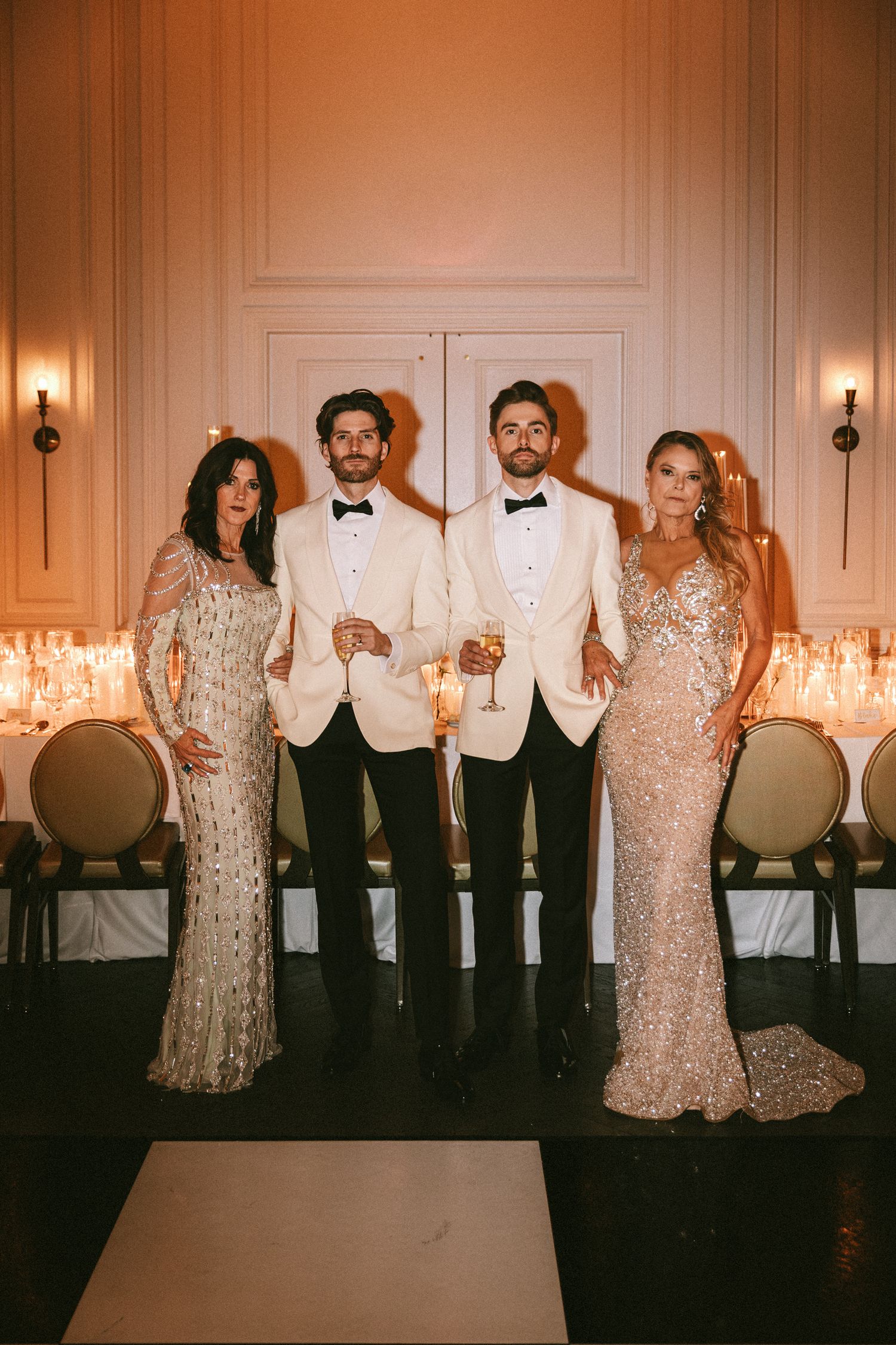 Two grooms in white and black tuxedos posing with family members in sequined gowns.