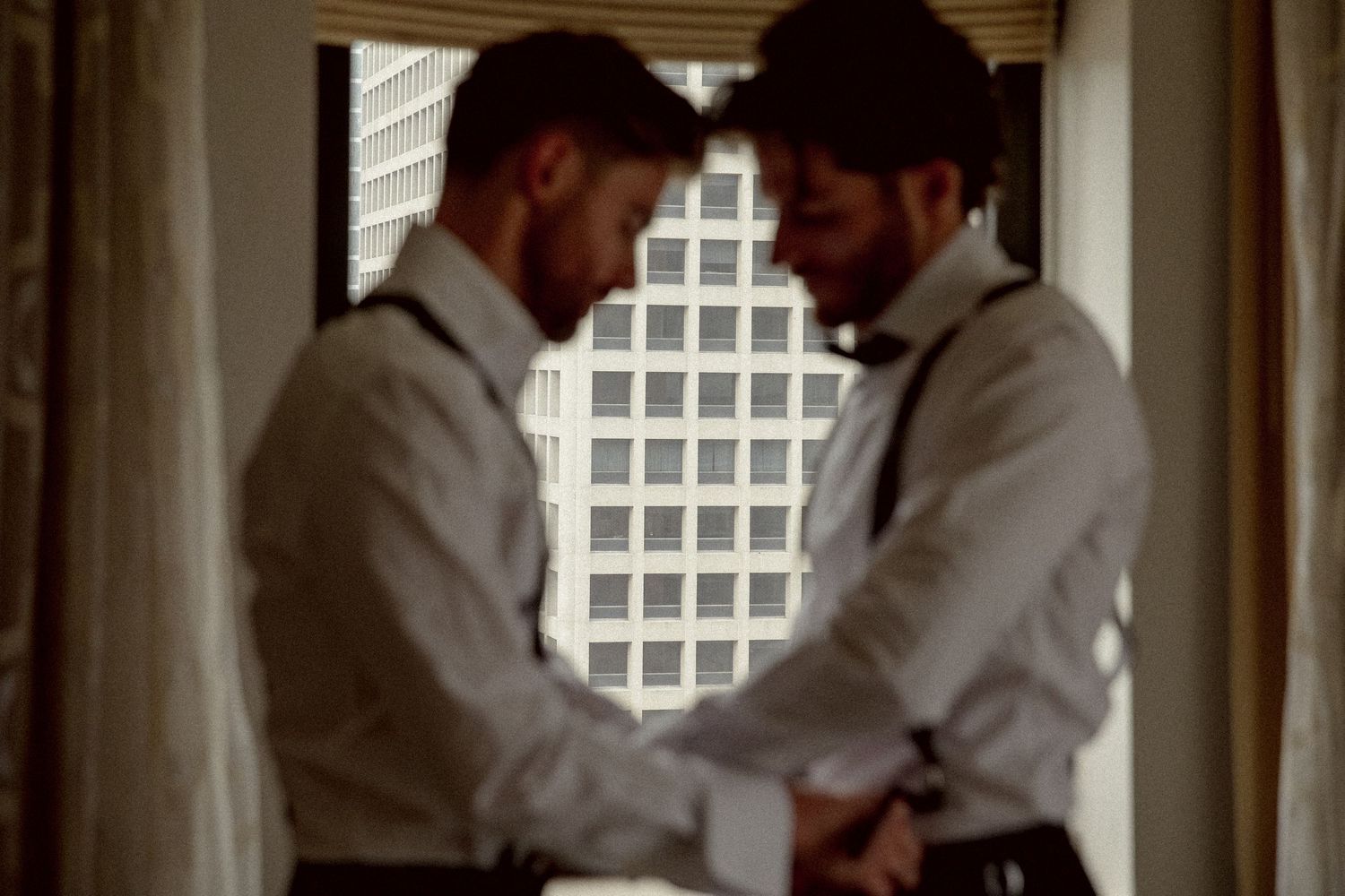 Two grooms in white shirts and suspenders sharing an intimate moment by a hotel window before wedding.