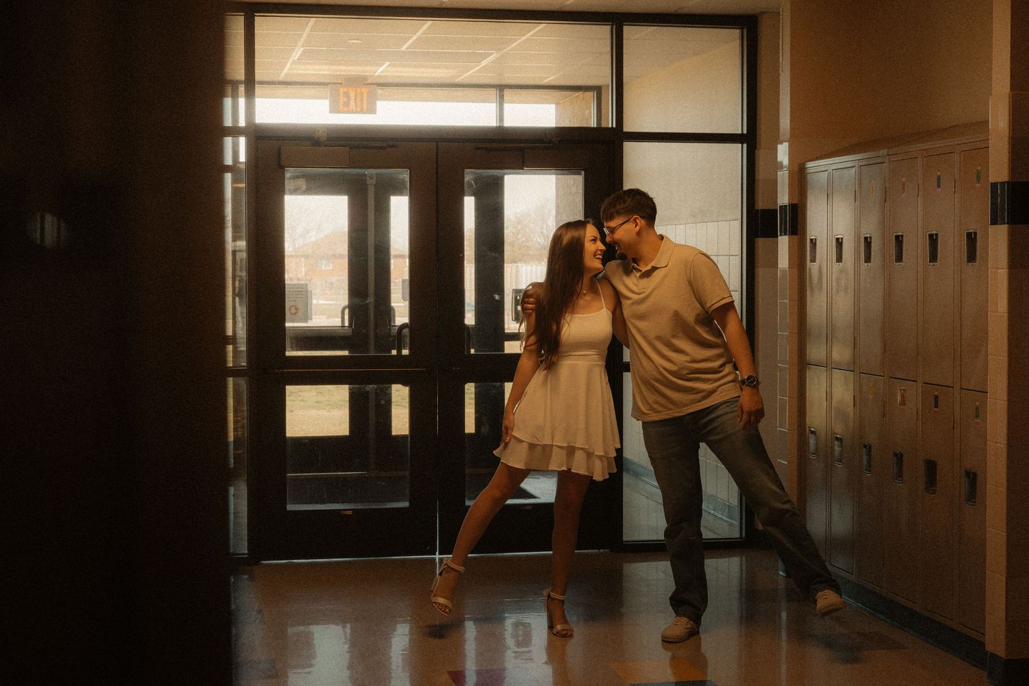Couple sharing a romantic moment in a school hallway near lockers and glass doors in warm moody lighting.