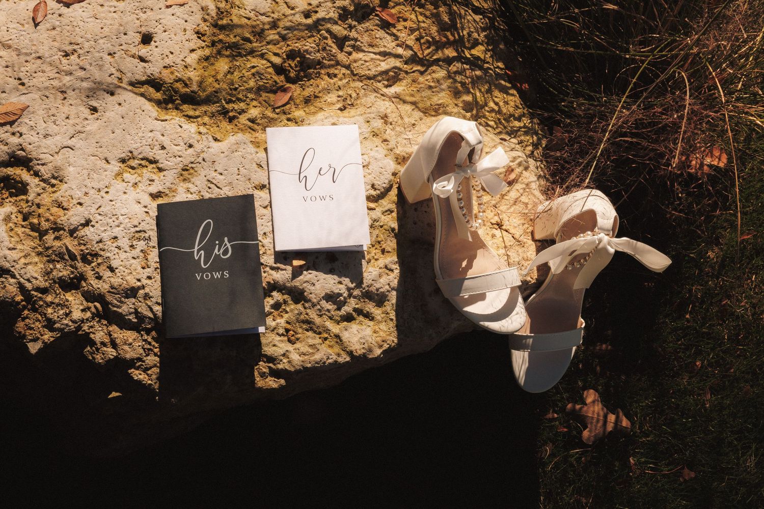 White bridal heels beside his and hers vow books on a rustic rock surface, perfect for wedding details.