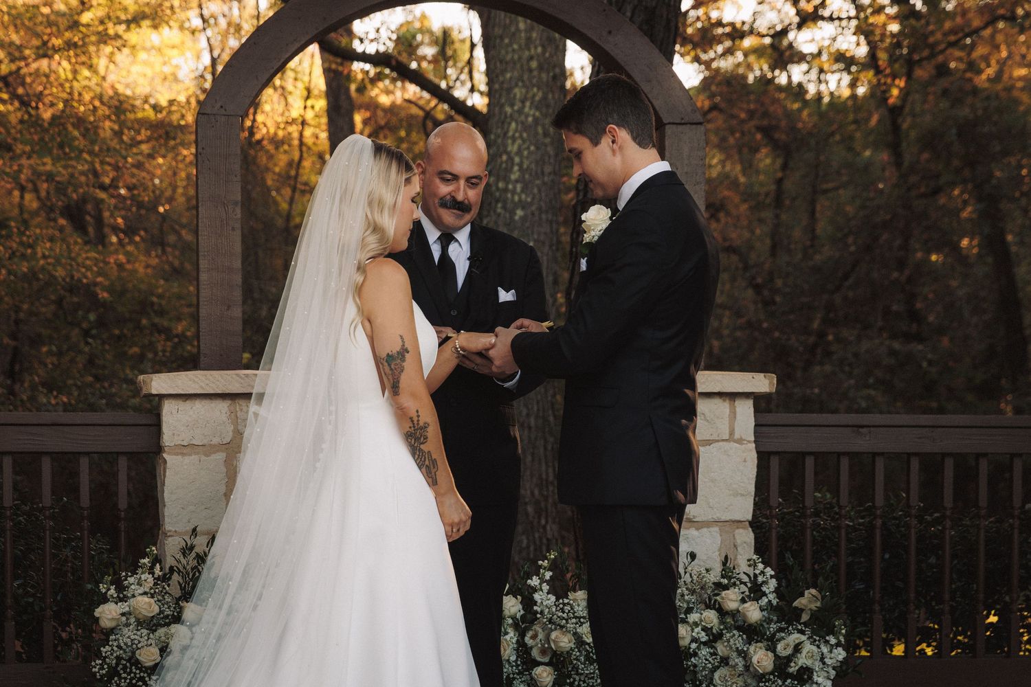 Bride and groom exchange vows under wooden arch with white floral arrangements at outdoor wedding ceremony.