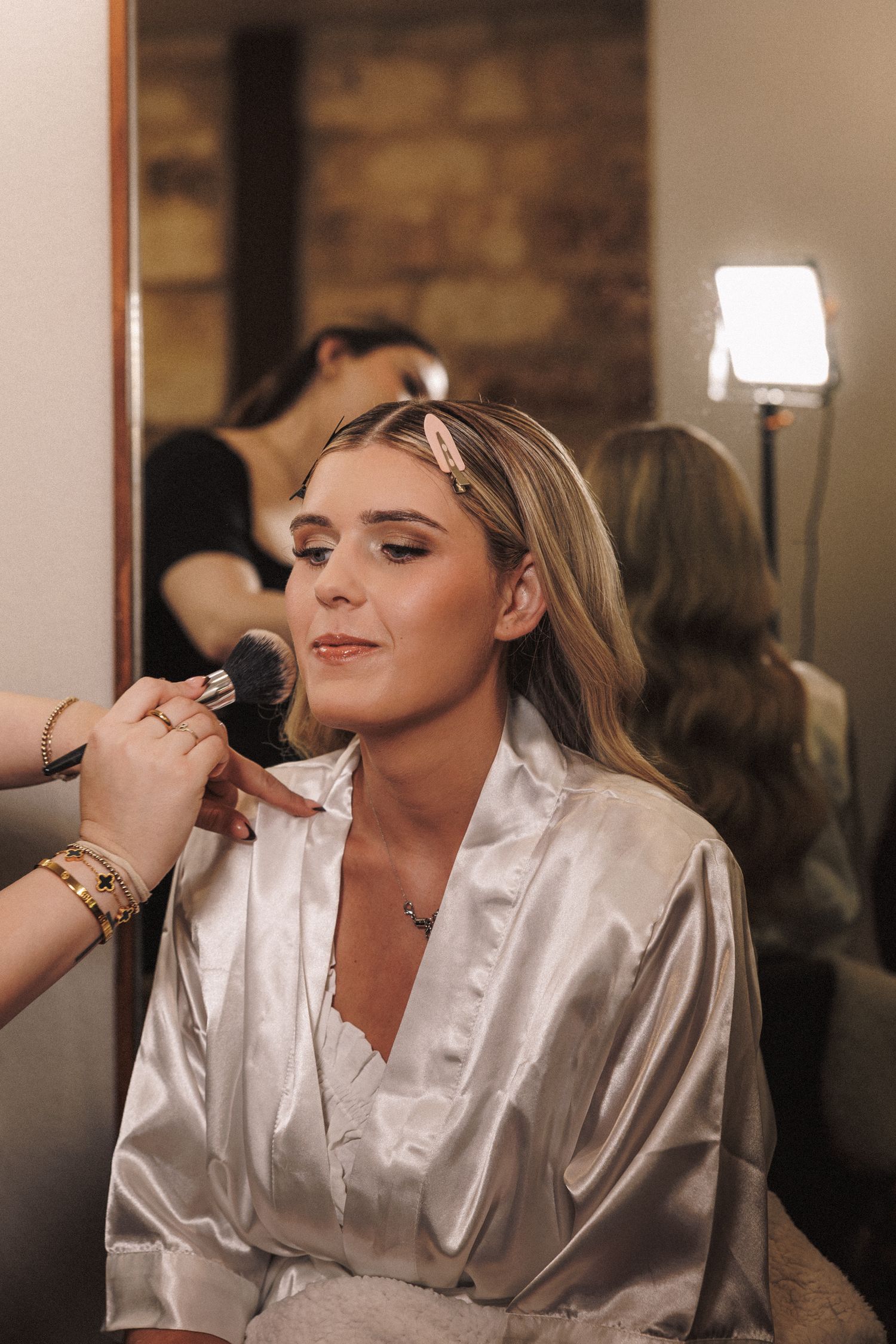 Blonde bride in satin robe getting makeup applied with a brush in a softly lit dressing room.