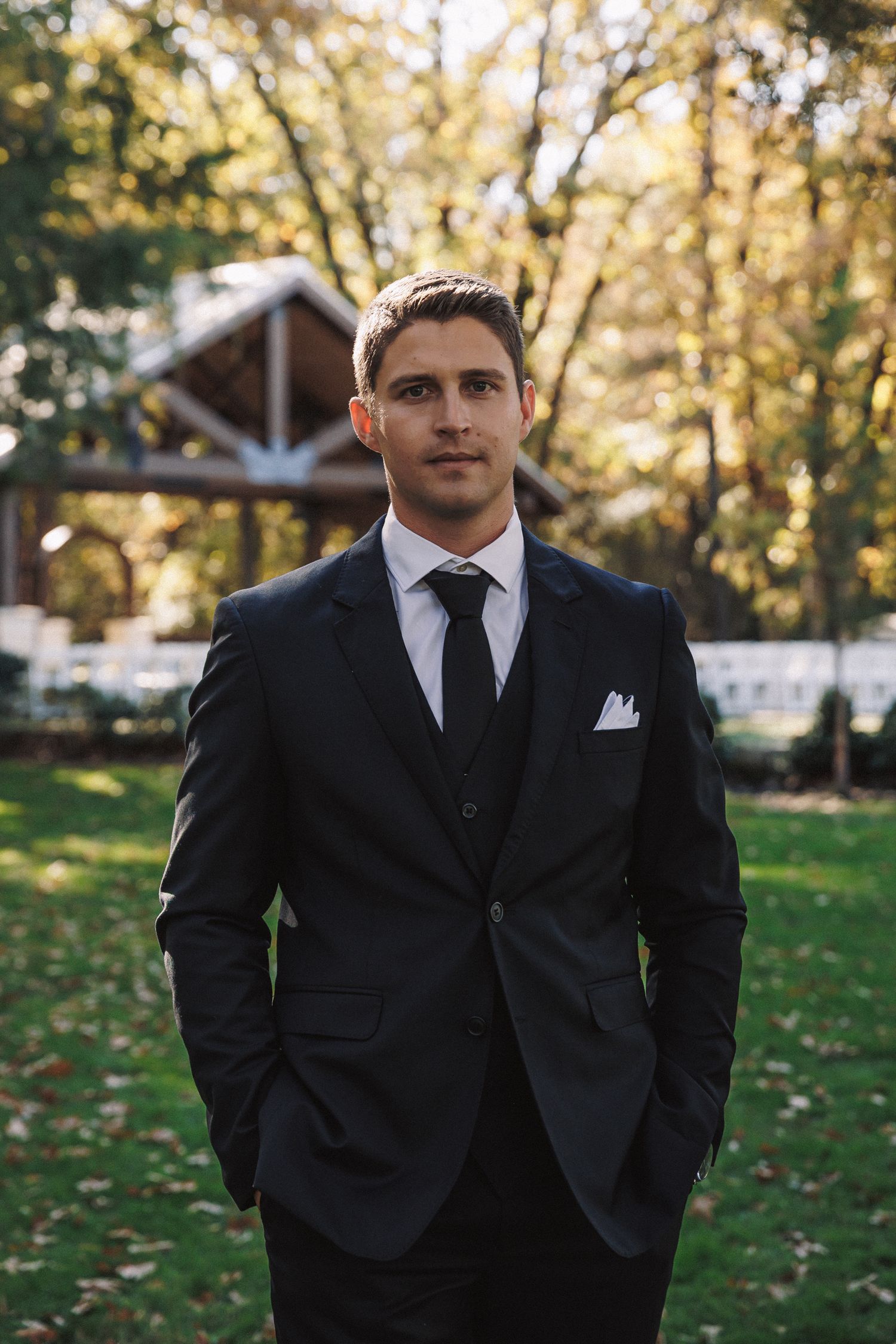 Handsome groom in black three-piece suit with tie and white pocket square standing outdoors at venue.