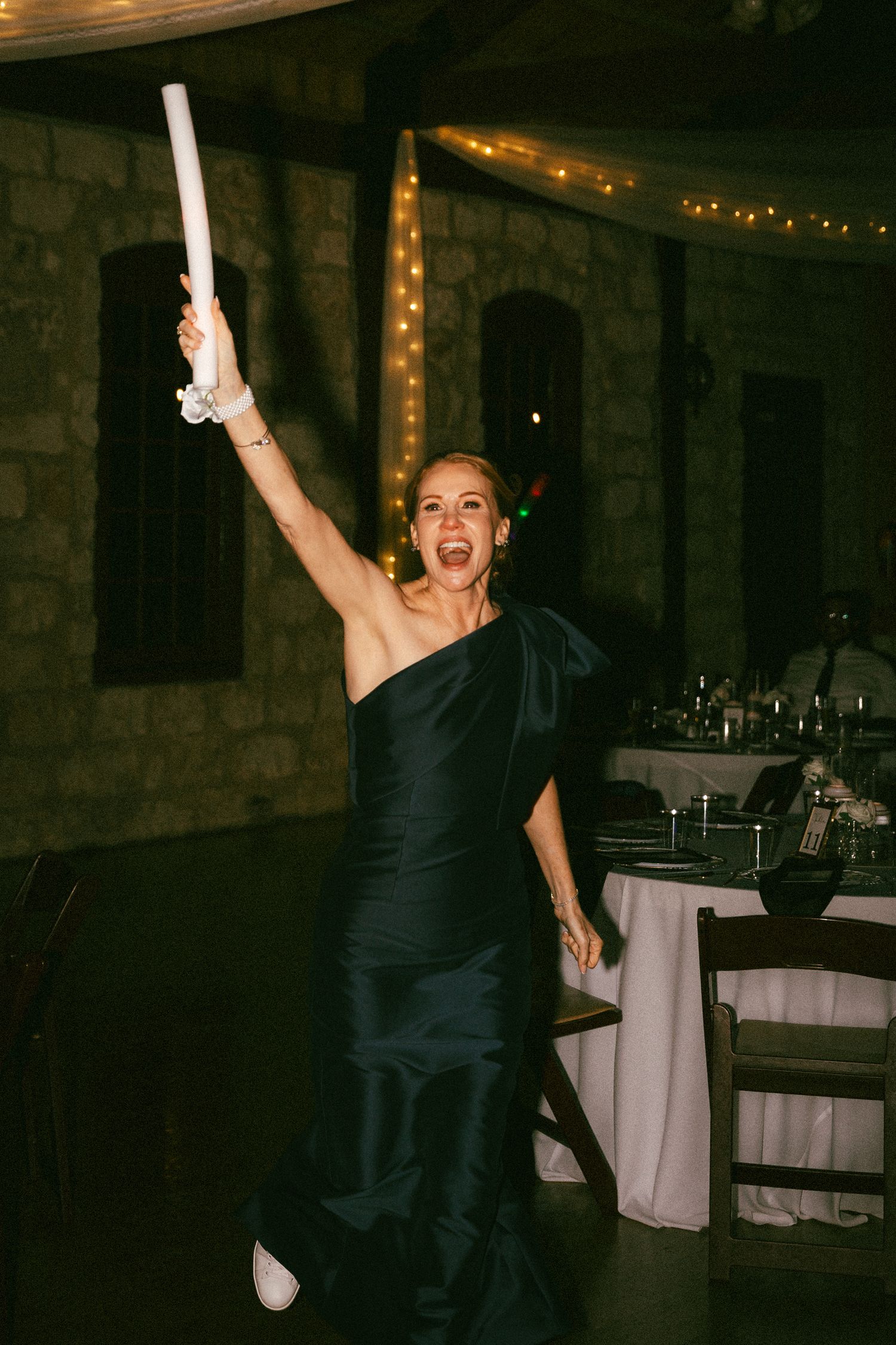 Woman in black one-shoulder gown celebrates at wedding reception raising a white cylinder in air.