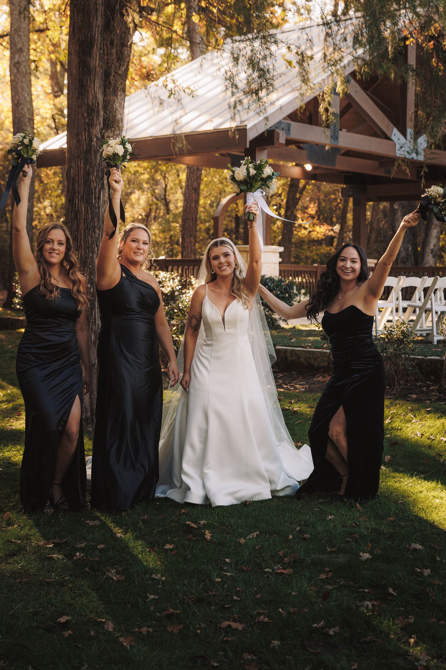 Bride and three bridesmaids in black gowns celebrate with raised bouquets at outdoor wedding.