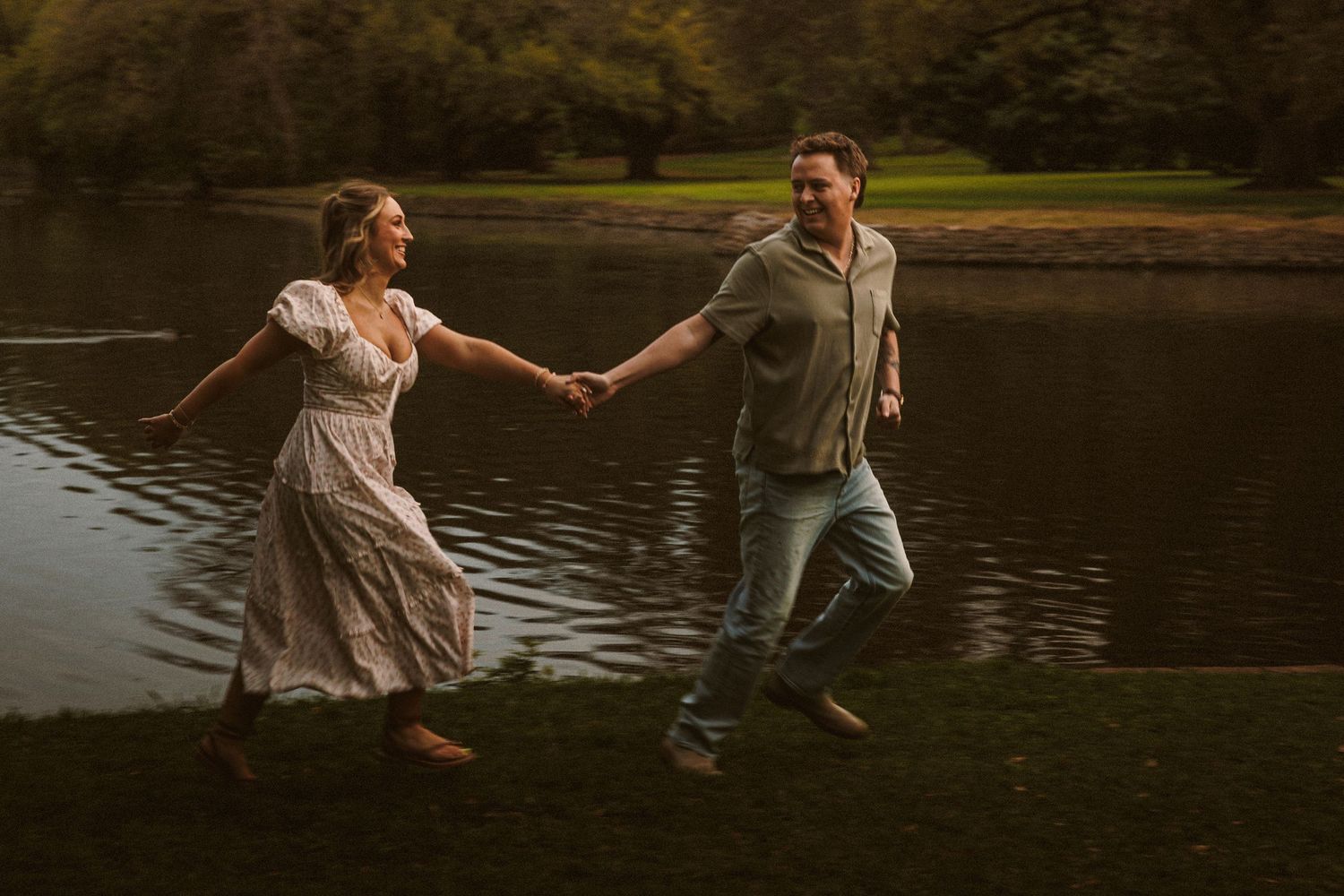 A joyful couple runs hand-in-hand along a serene lakeside park during golden hour.