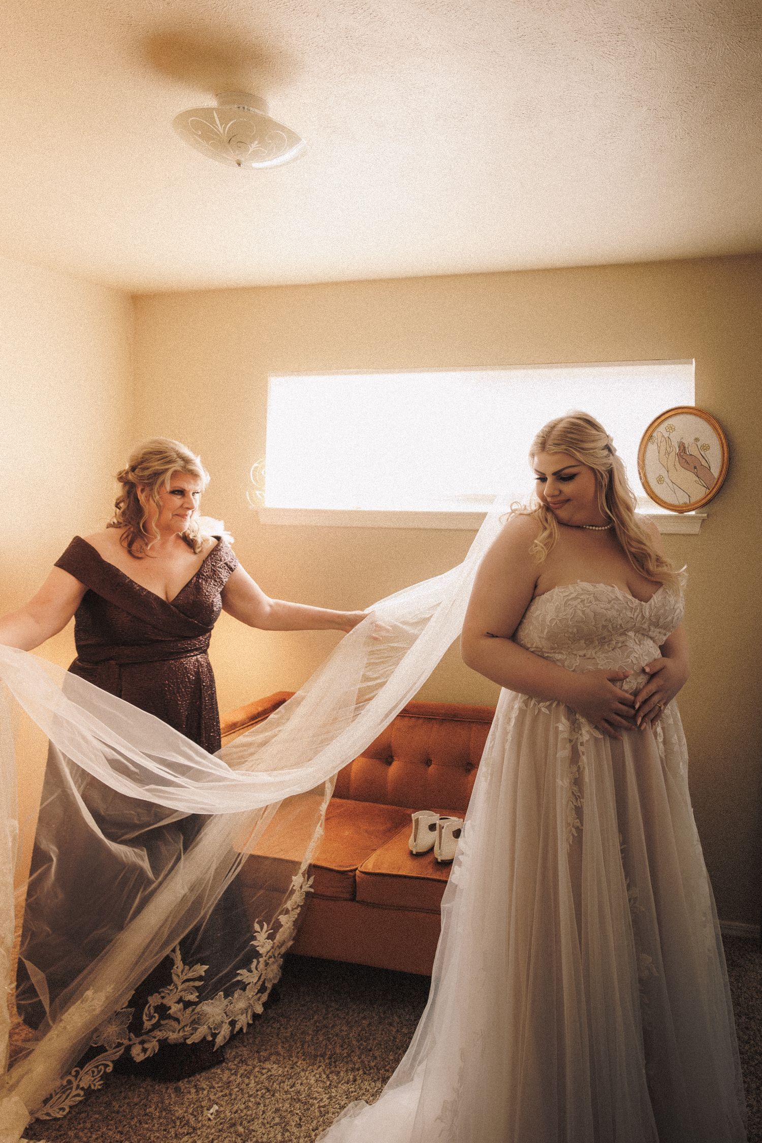 Mother helping bride with lace wedding veil in warm sunlit room before ceremony.