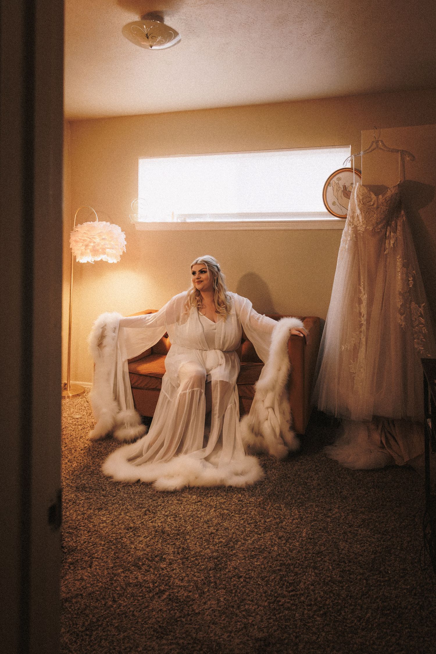 Bride in white fur-trimmed robe sitting in warmly lit room with wedding dress hanging nearby.