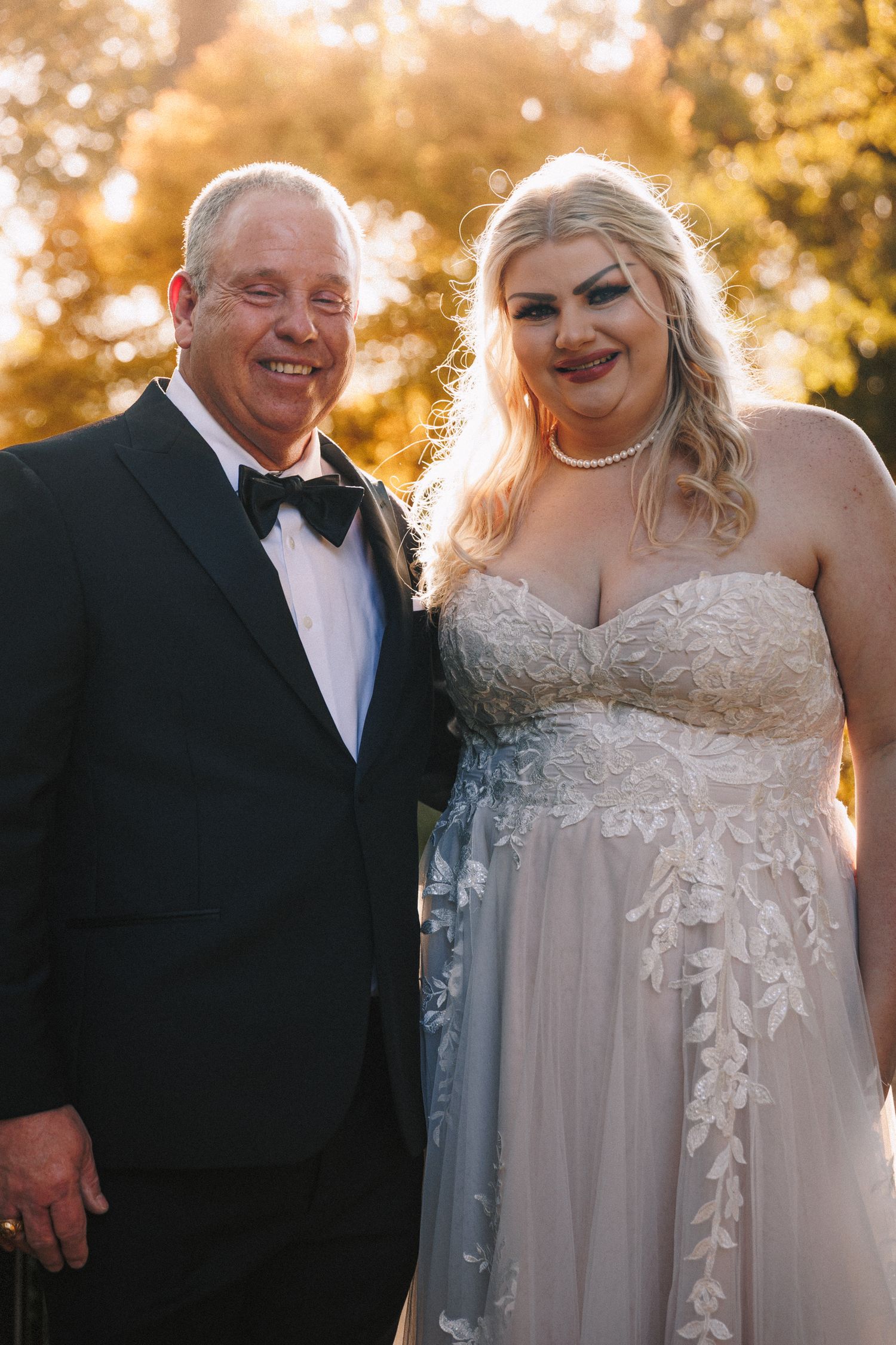 Bride in floral lace gown poses with smiling father in tuxedo during golden hour outdoor wedding.