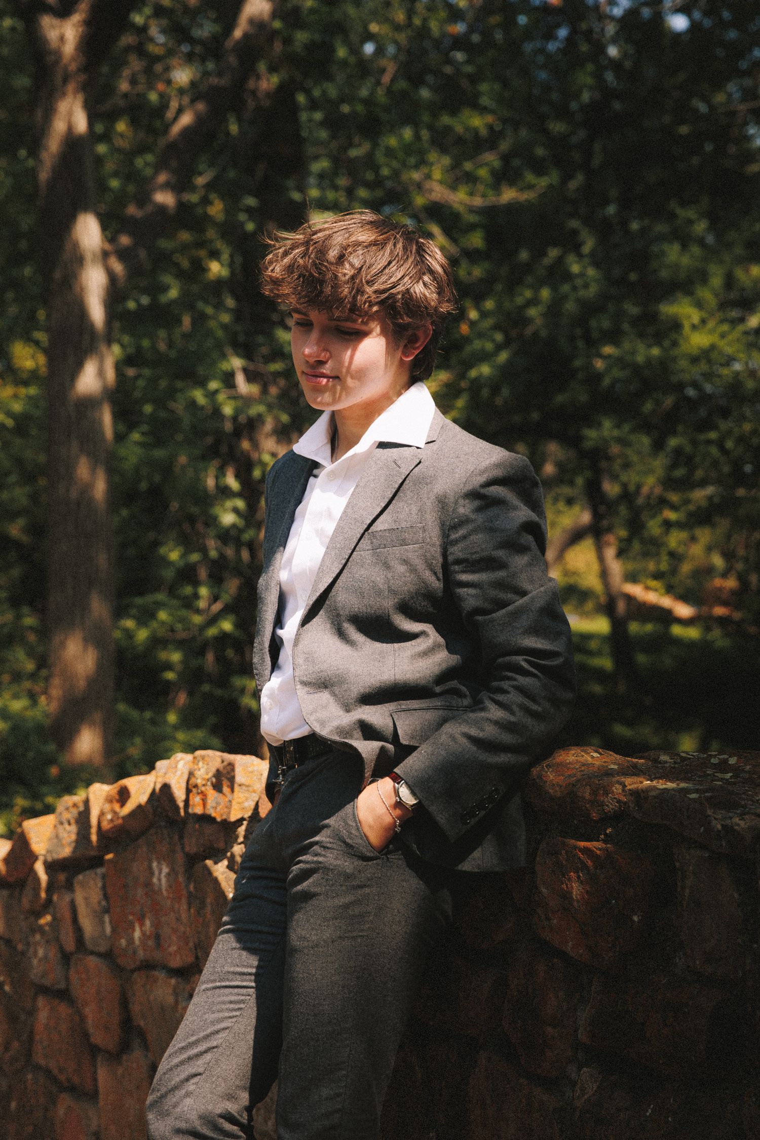 Person in a gray suit leans against a stone wall in a sunlit garden setting.