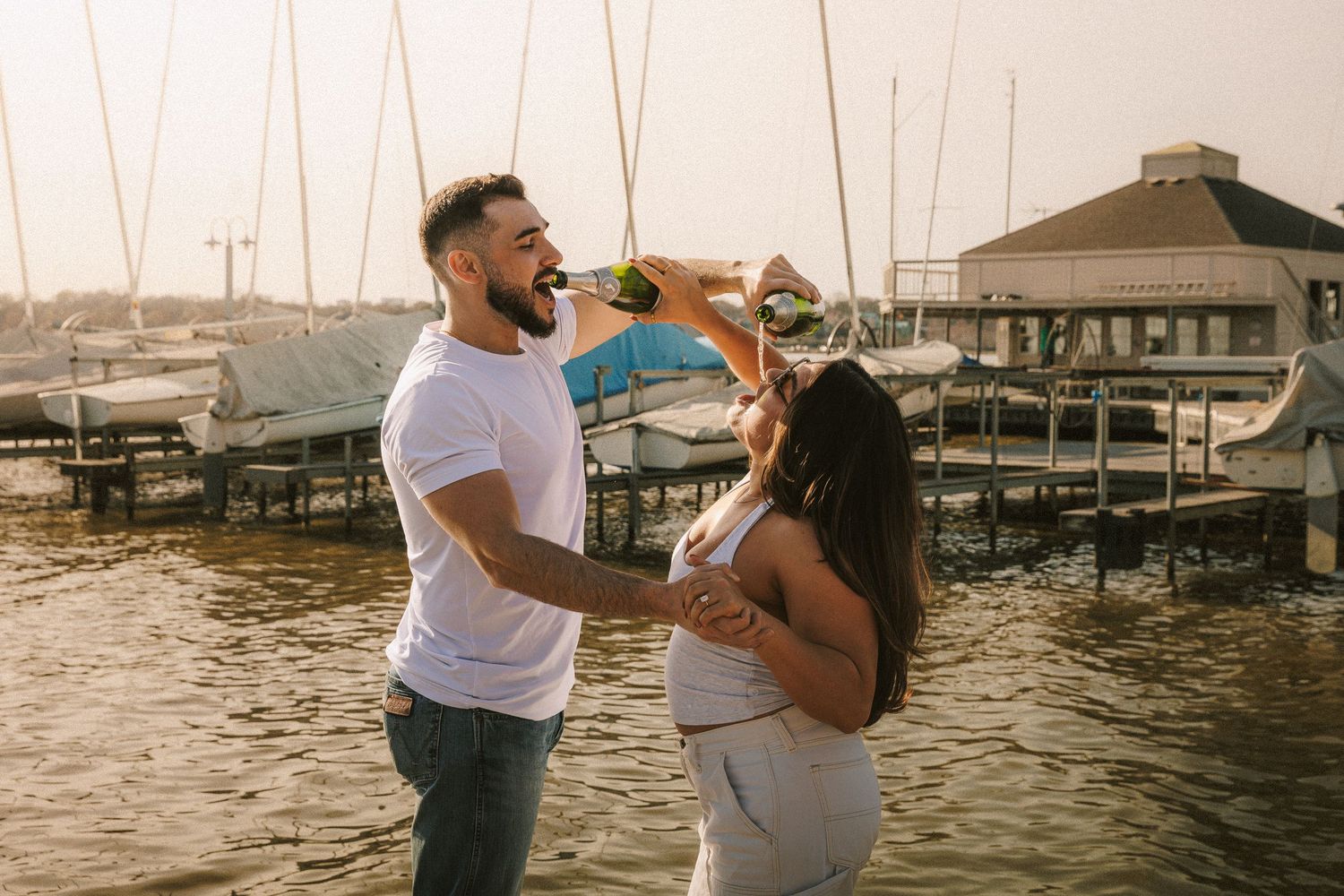 A couple shares a joyful moment at sunset by a marina with boats and a dock house in the background.