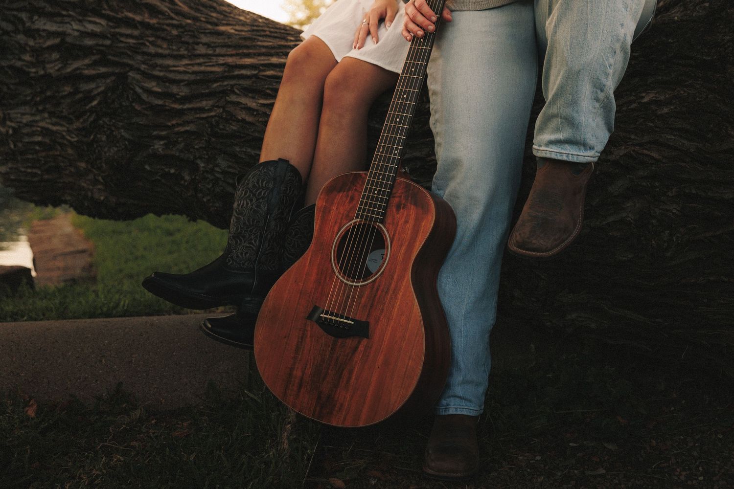 A person wearing jeans holds an acoustic guitar while standing in front of a dark textured background.