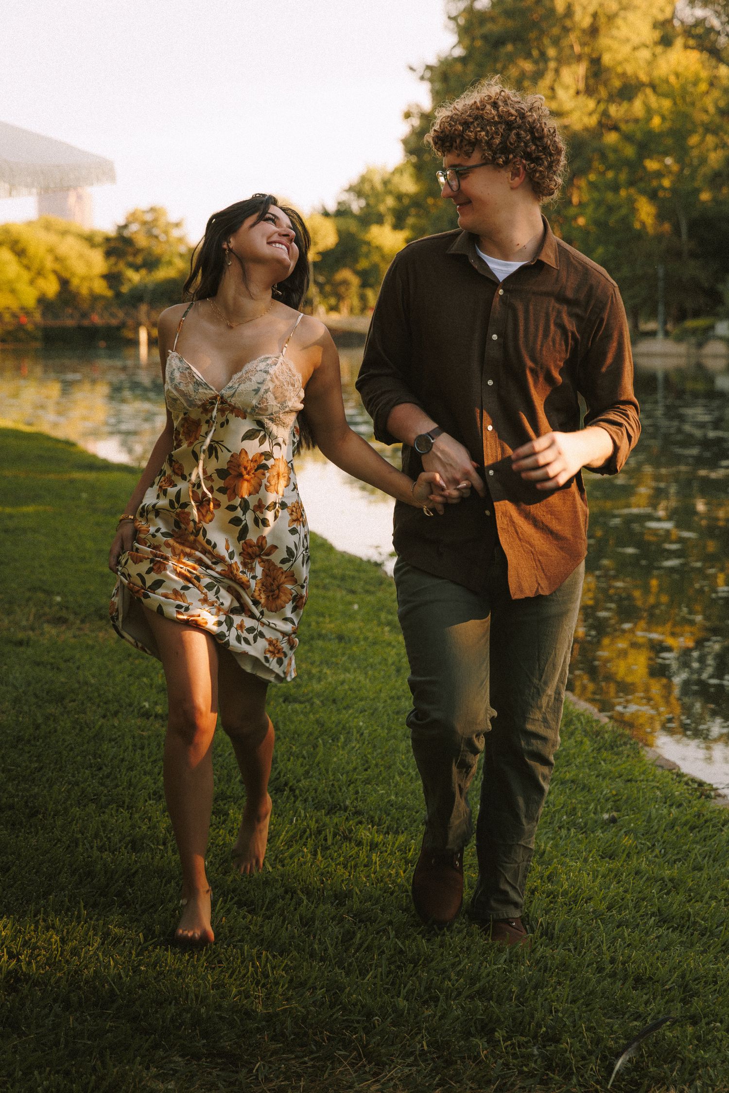 Couple walking together on grassy path by water during warm sunset lighting.