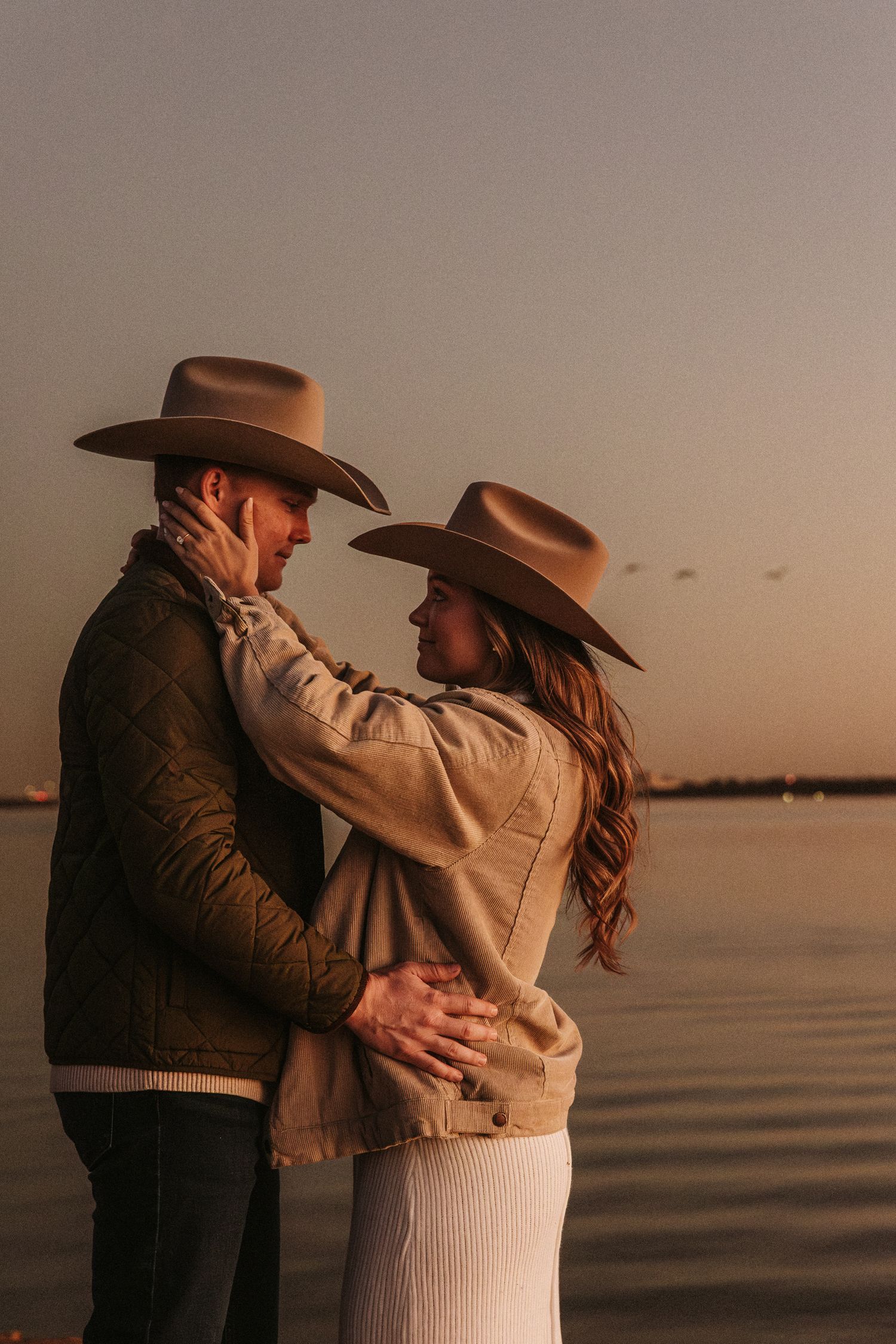 Couple in western attire sharing intimate moments by the water during sunset.