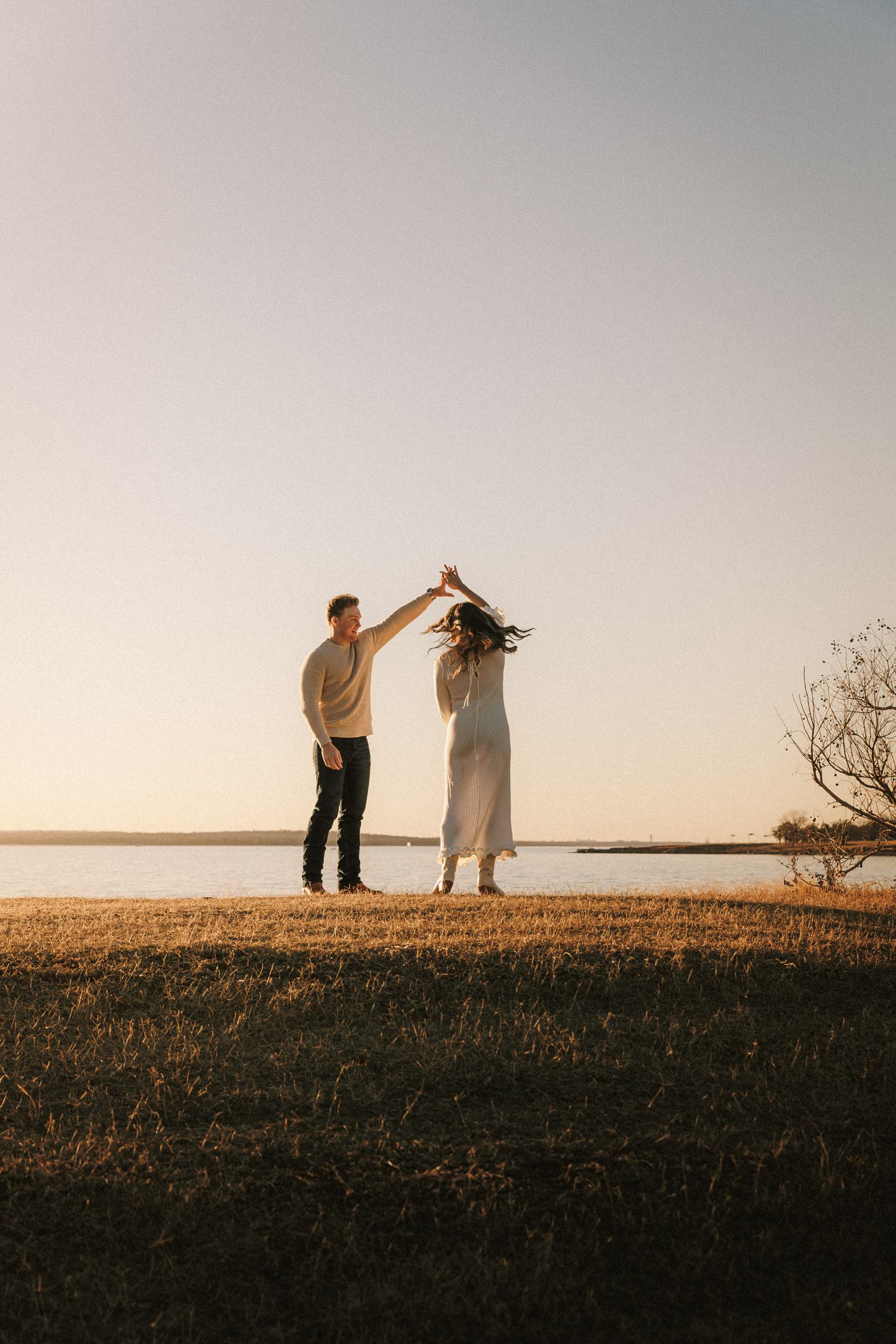 A couple dances together on a hilltop against a golden sunset sky and lake backdrop.