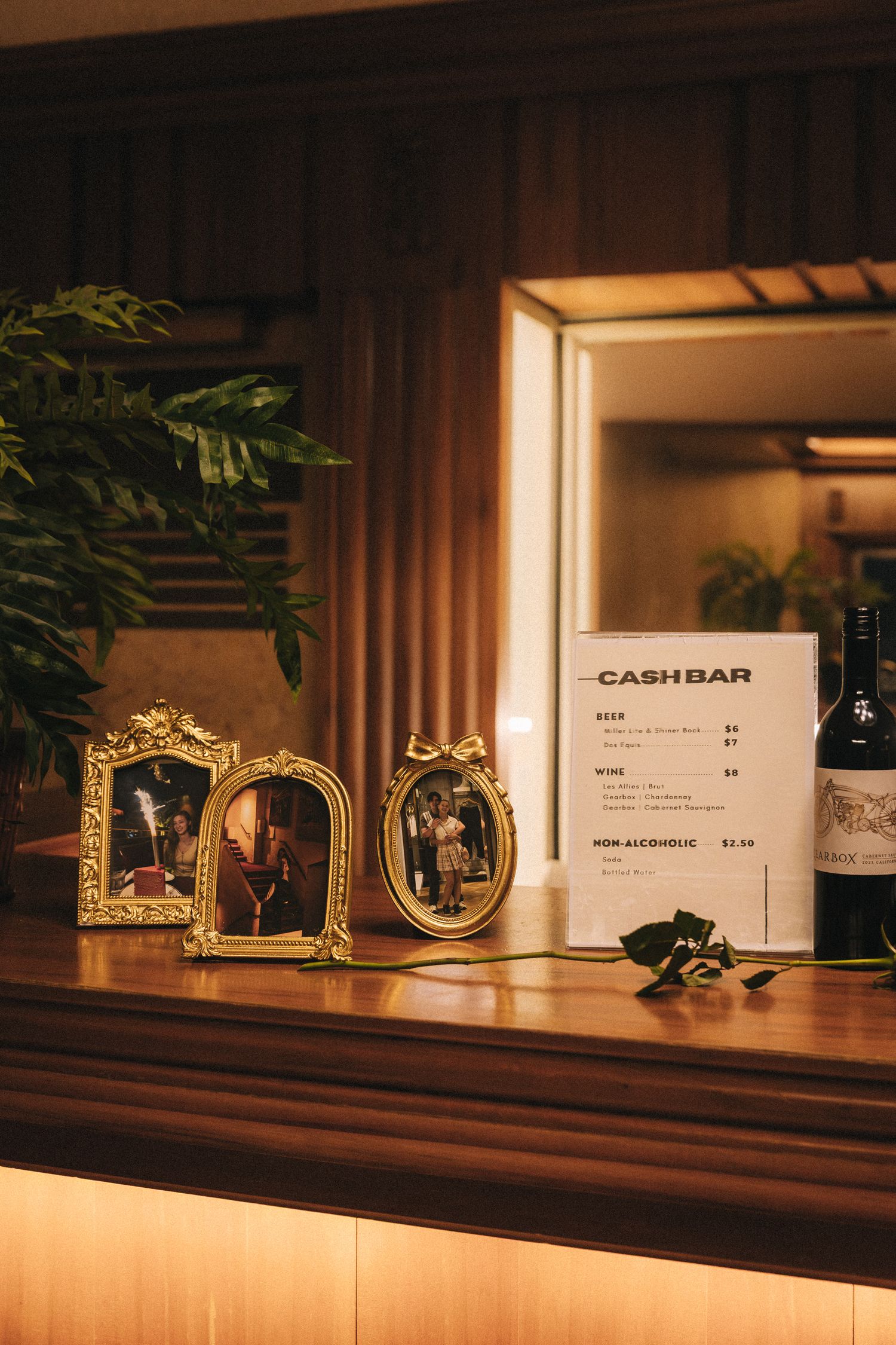 Golden-framed photos and a cash bar menu sign on a warmly lit wooden counter at an elegant event venue.