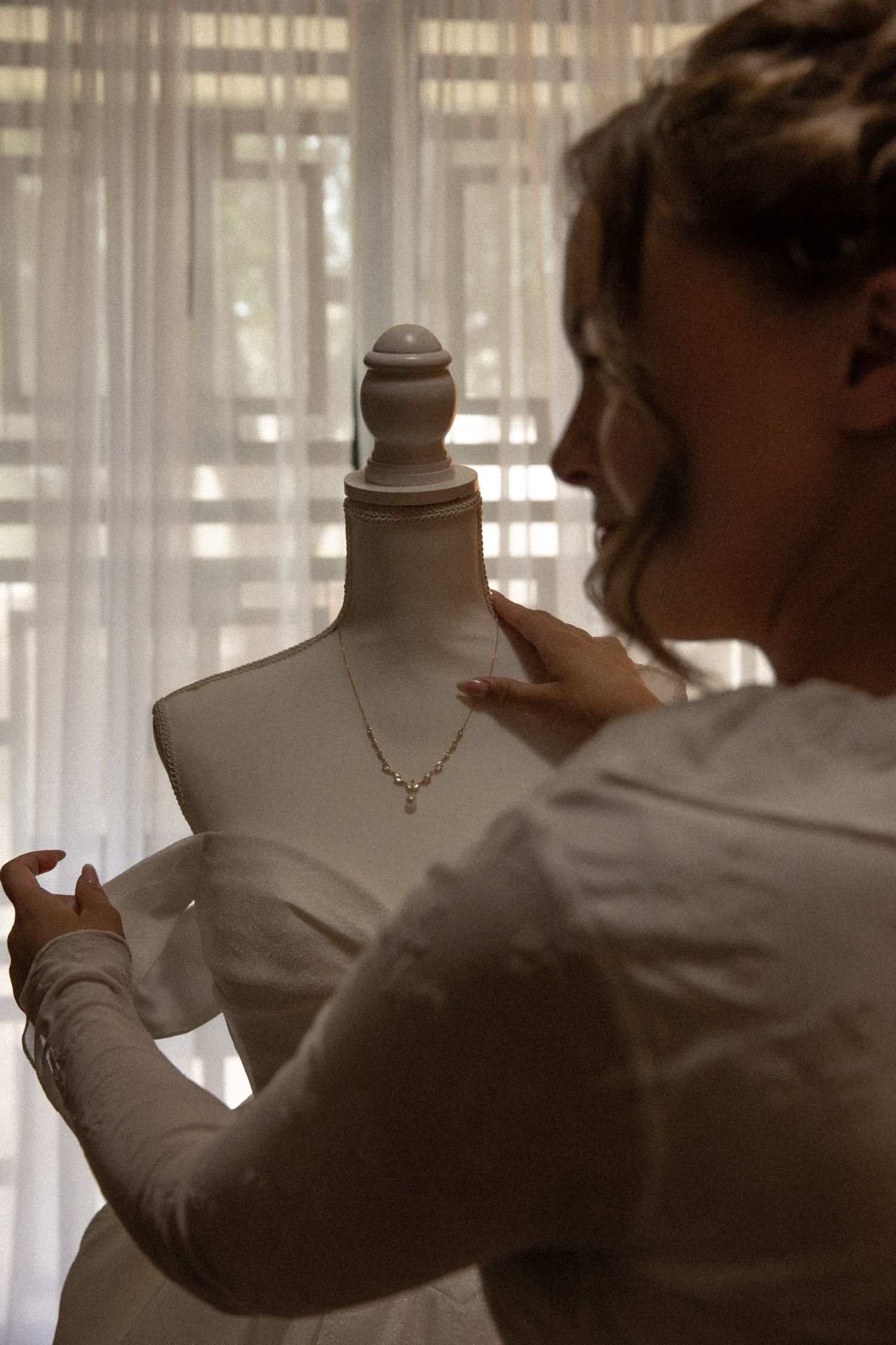Bride in white dress adjusting a delicate necklace on a vintage mannequin bust near a curtained window.