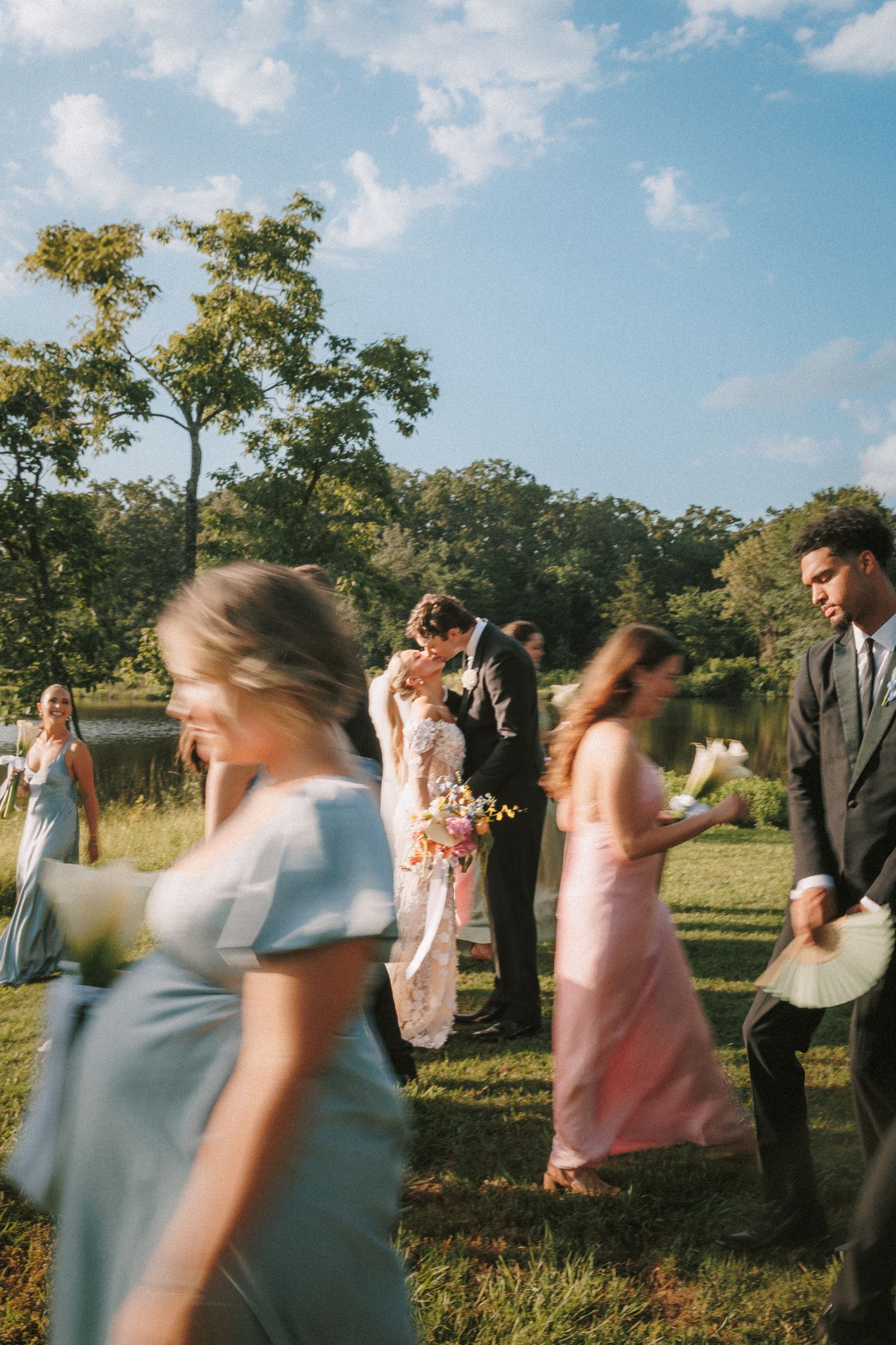 Wedding guests mingle during an outdoor summer celebration with trees and a lake in the background.