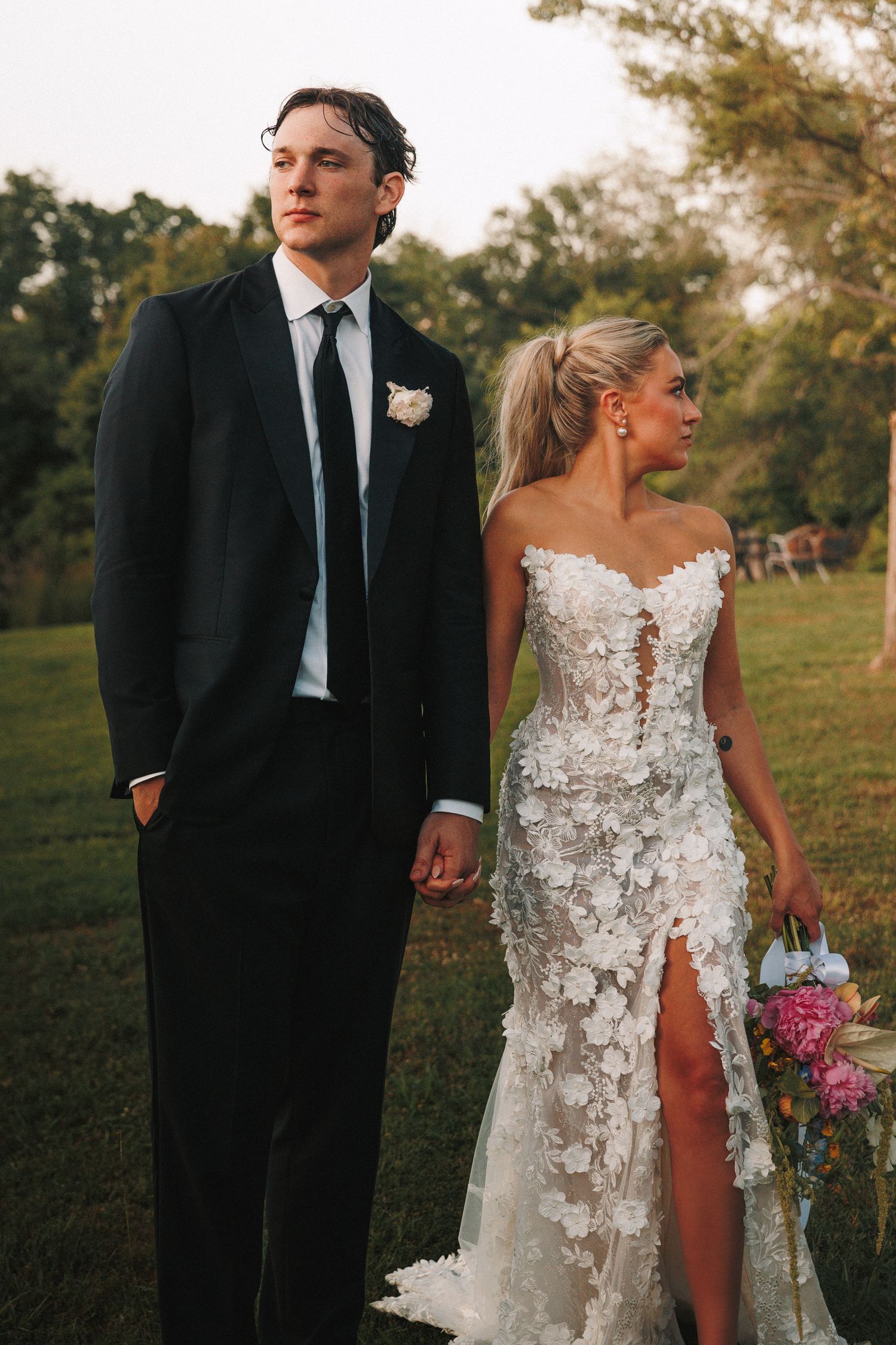 A newlywed couple stands in a garden setting, with the bride wearing an elegant lace gown with a side slit.
