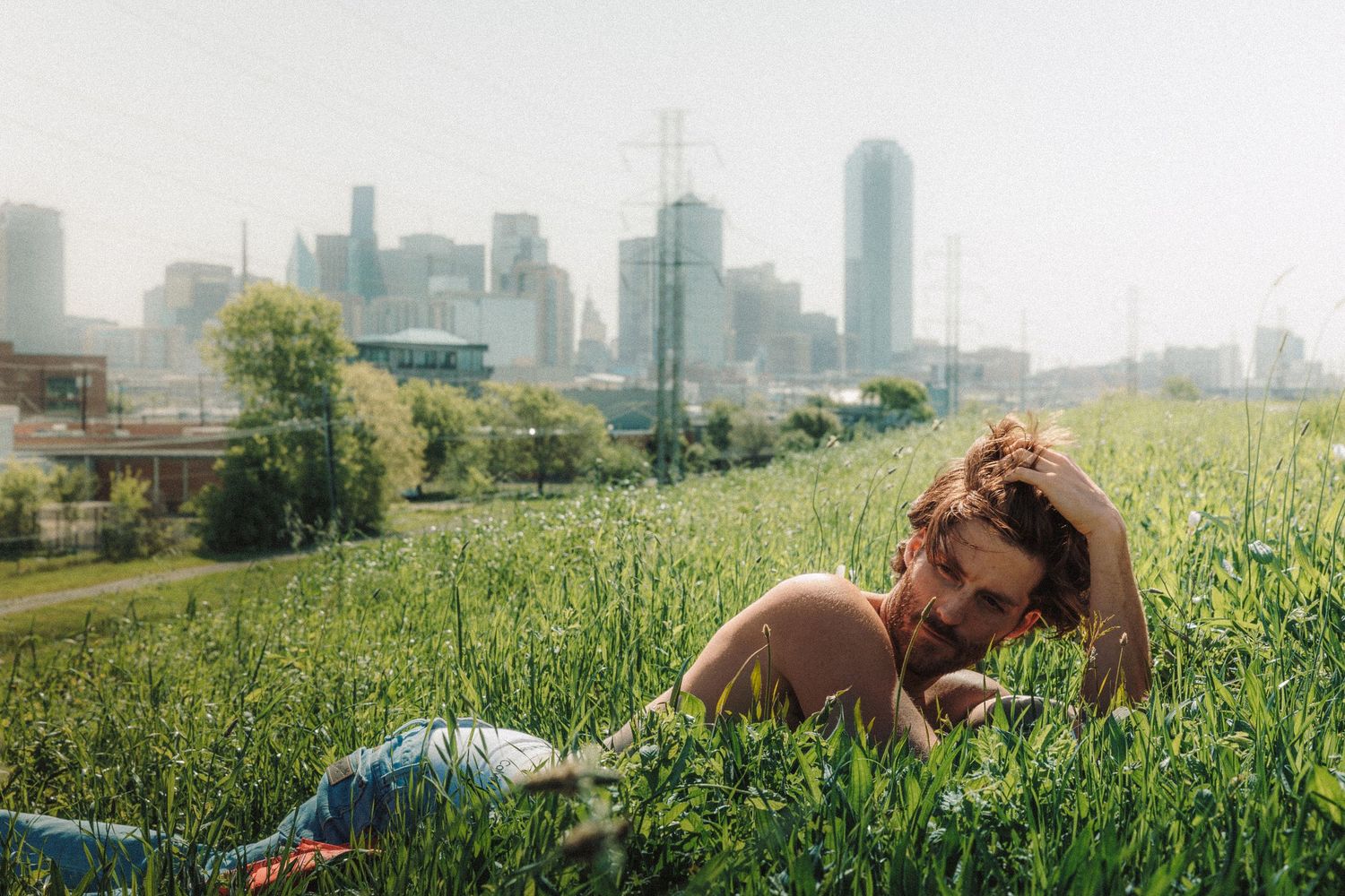 Someone relaxes in green grass on a hill with downtown city skyline visible in the background on a hazy day.
