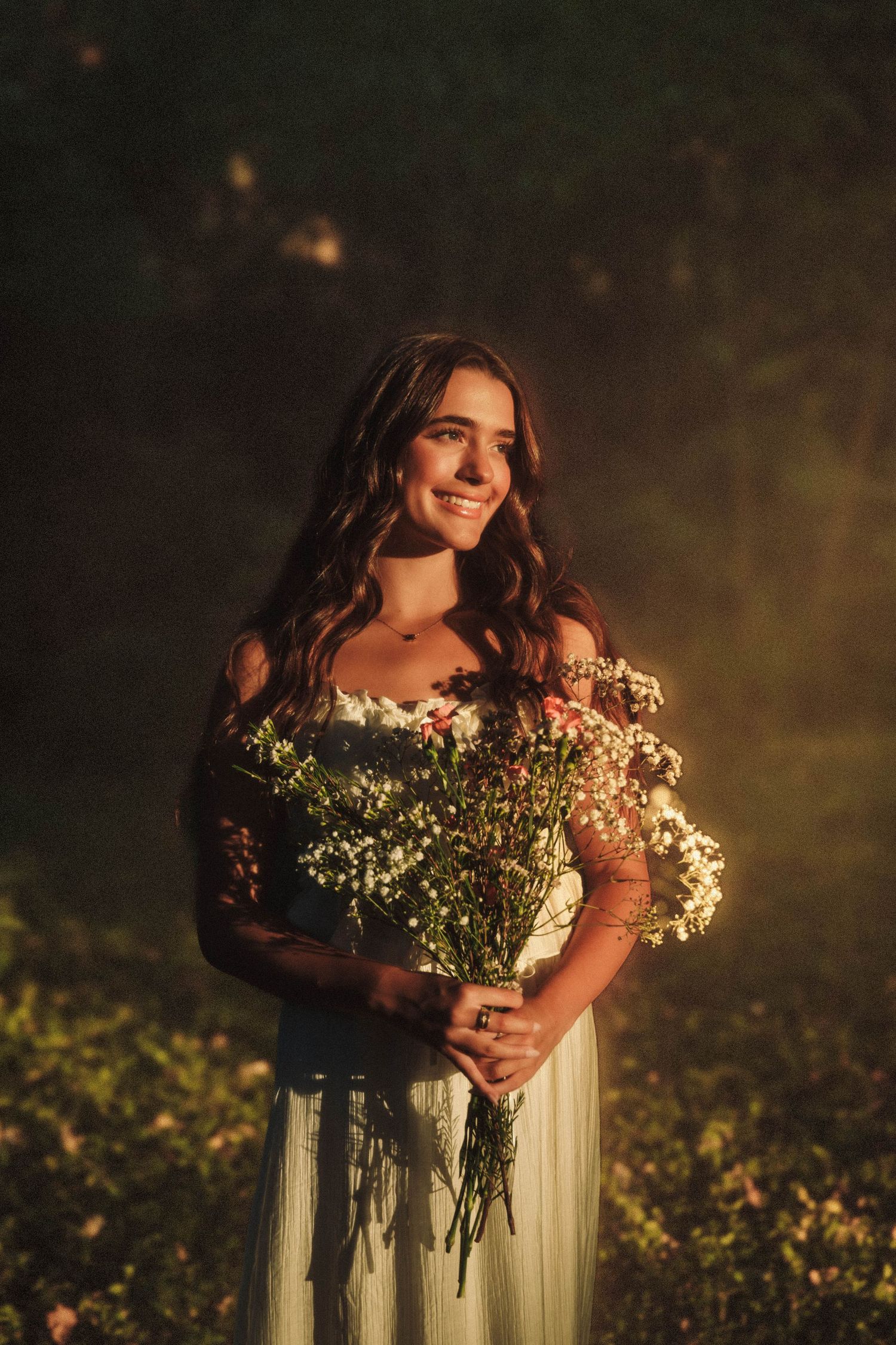 A romantic outdoor portrait of someone holding delicate white wildflowers while wearing a flowing white dress in moody lighting.