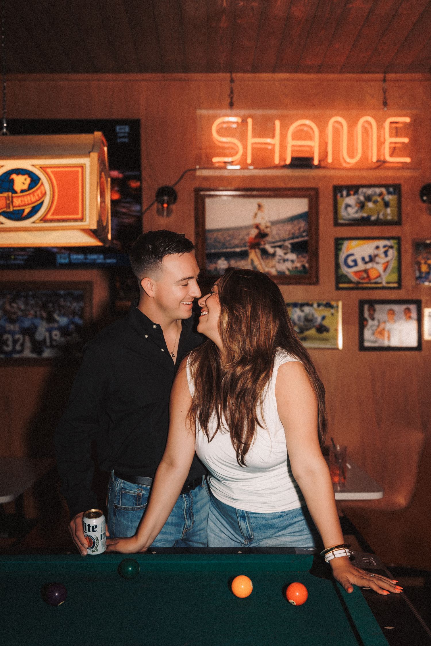 Two people play pool in a dimly lit bar with neon signs and vintage decor on the walls.