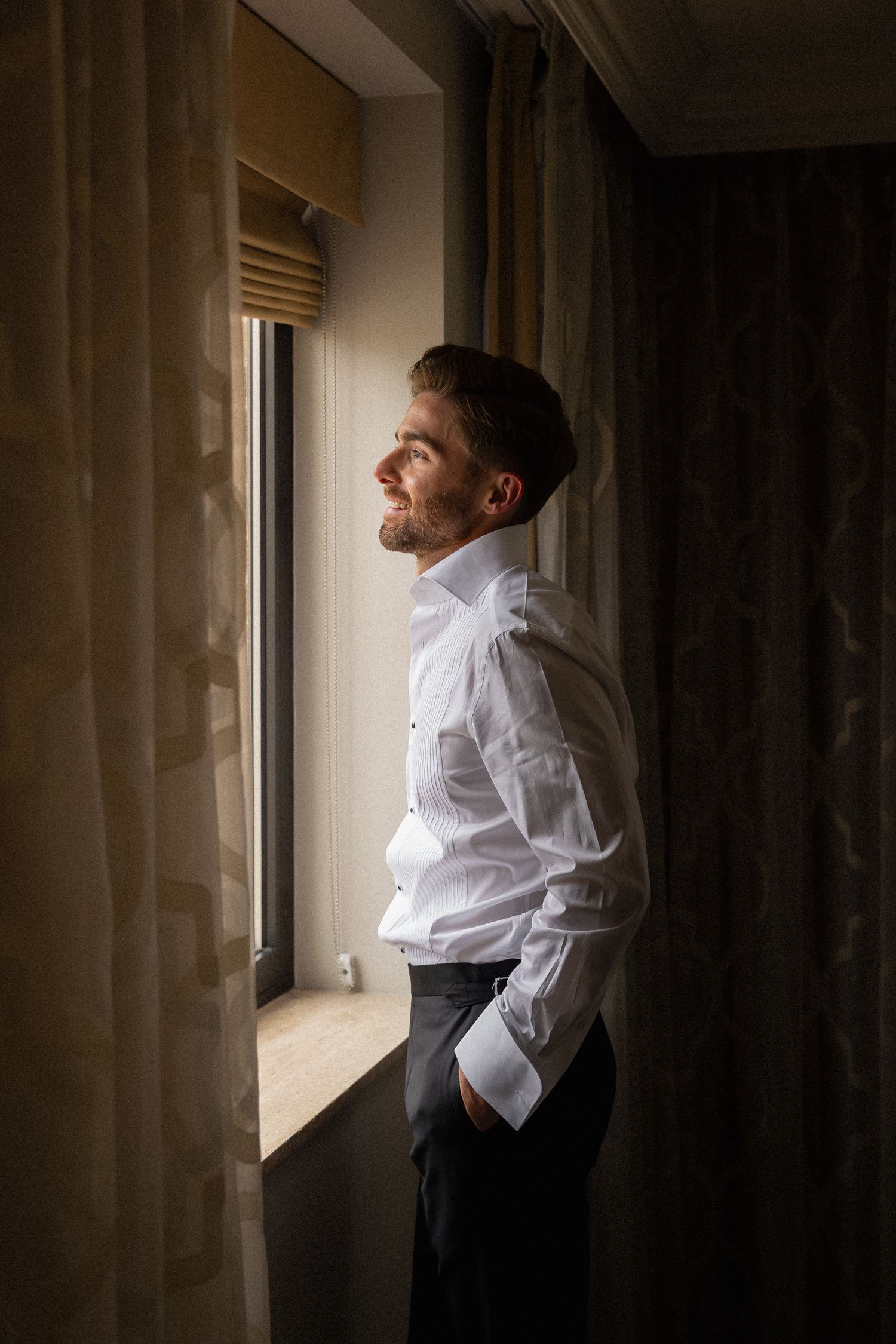 Groom in white dress shirt gazing out window with natural light, smiling in anticipation on wedding day.