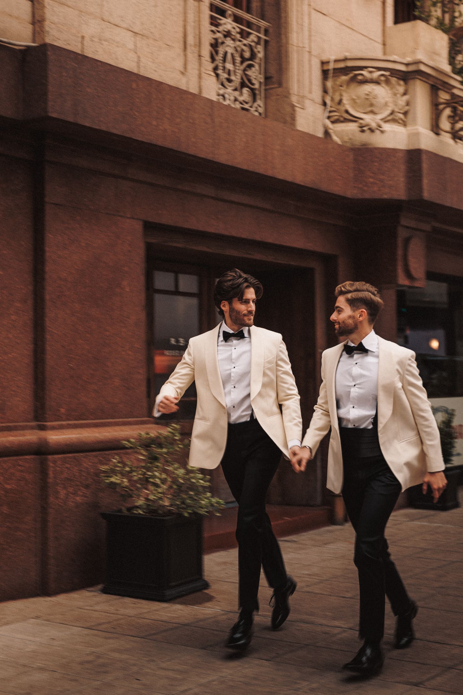 Two grooms in matching cream blazers holding hands walking outside elegant building.