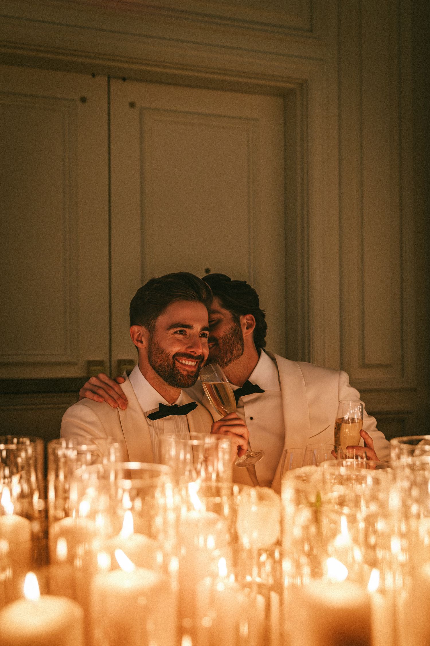 Two grooms share a joyful candlelit moment at wedding reception table surrounded by glowing candles.