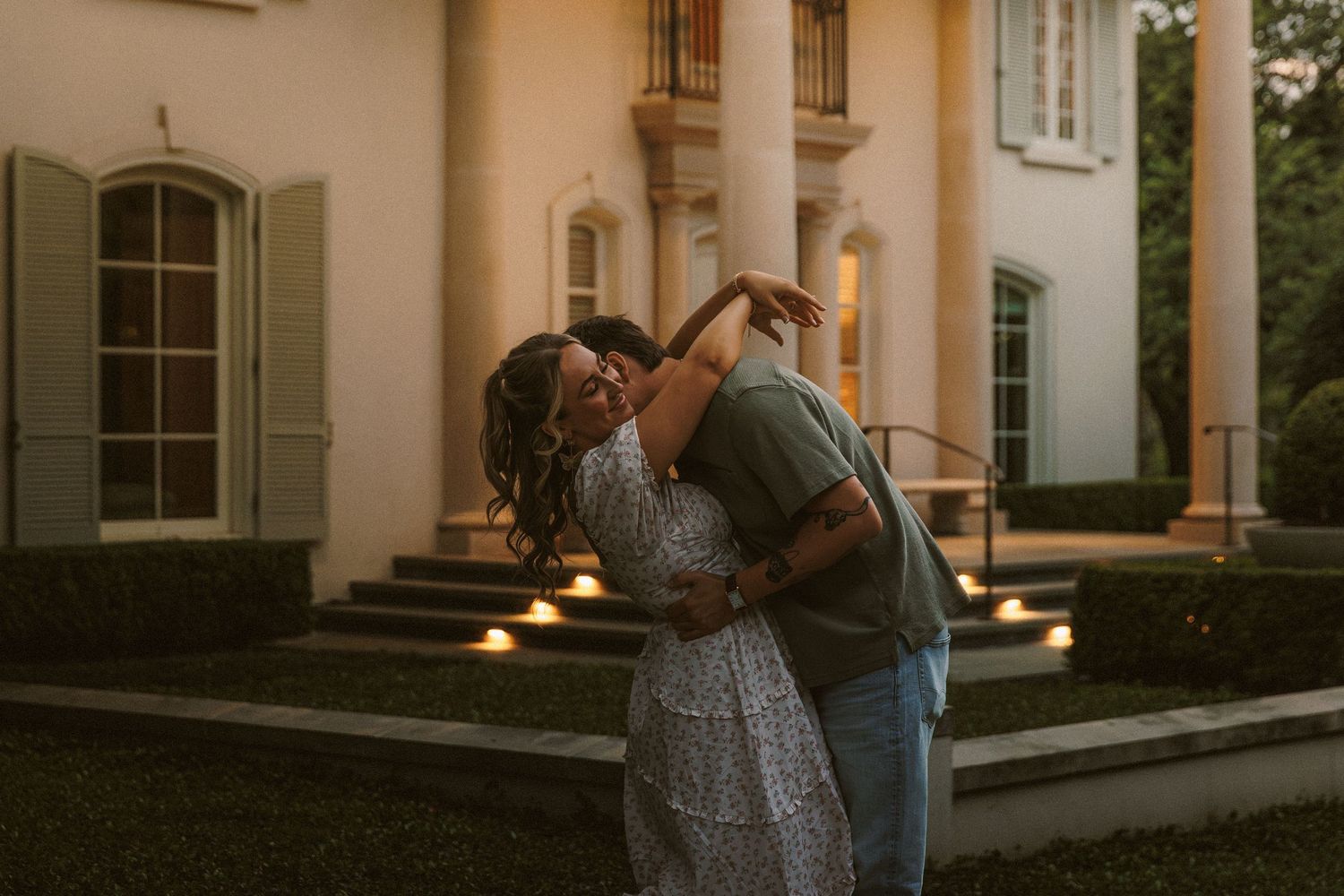 Couple embracing romantically at dusk outside elegant white colonial mansion with glowing candles.