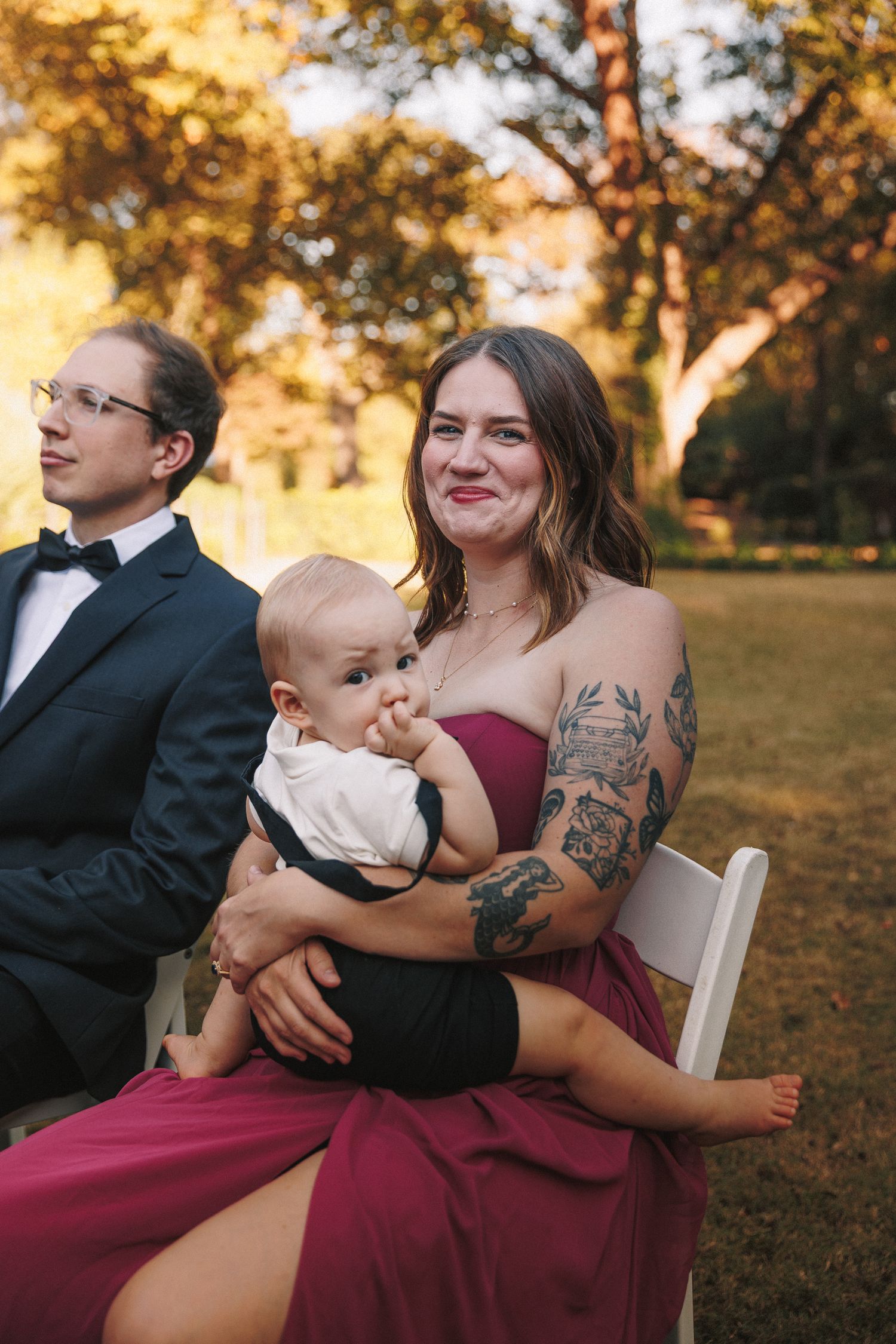 Wedding guest in burgundy gown holds infant while seated at outdoor autumn ceremony.