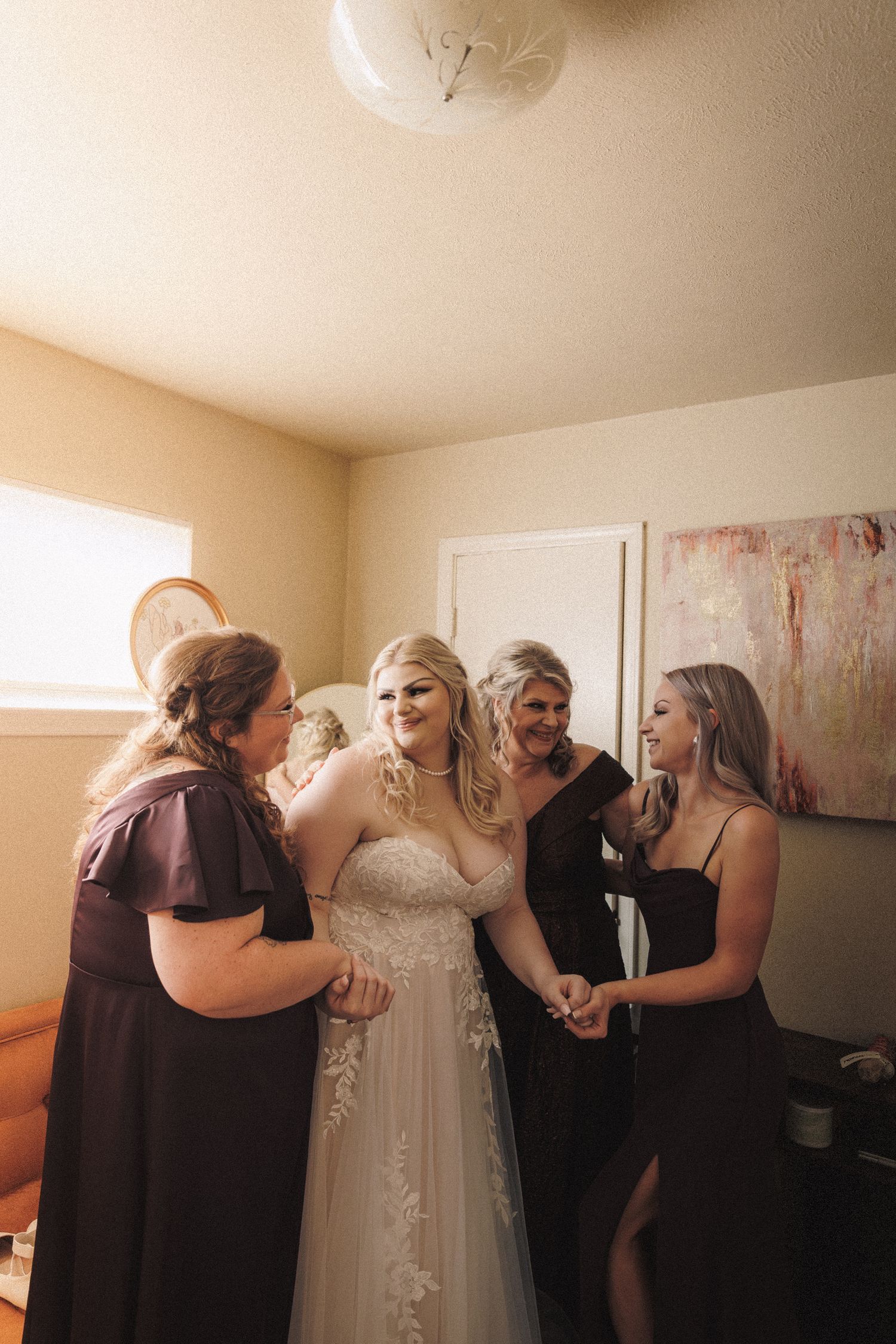 Bride in lace wedding gown laughing with bridesmaids in dark dresses during getting ready.