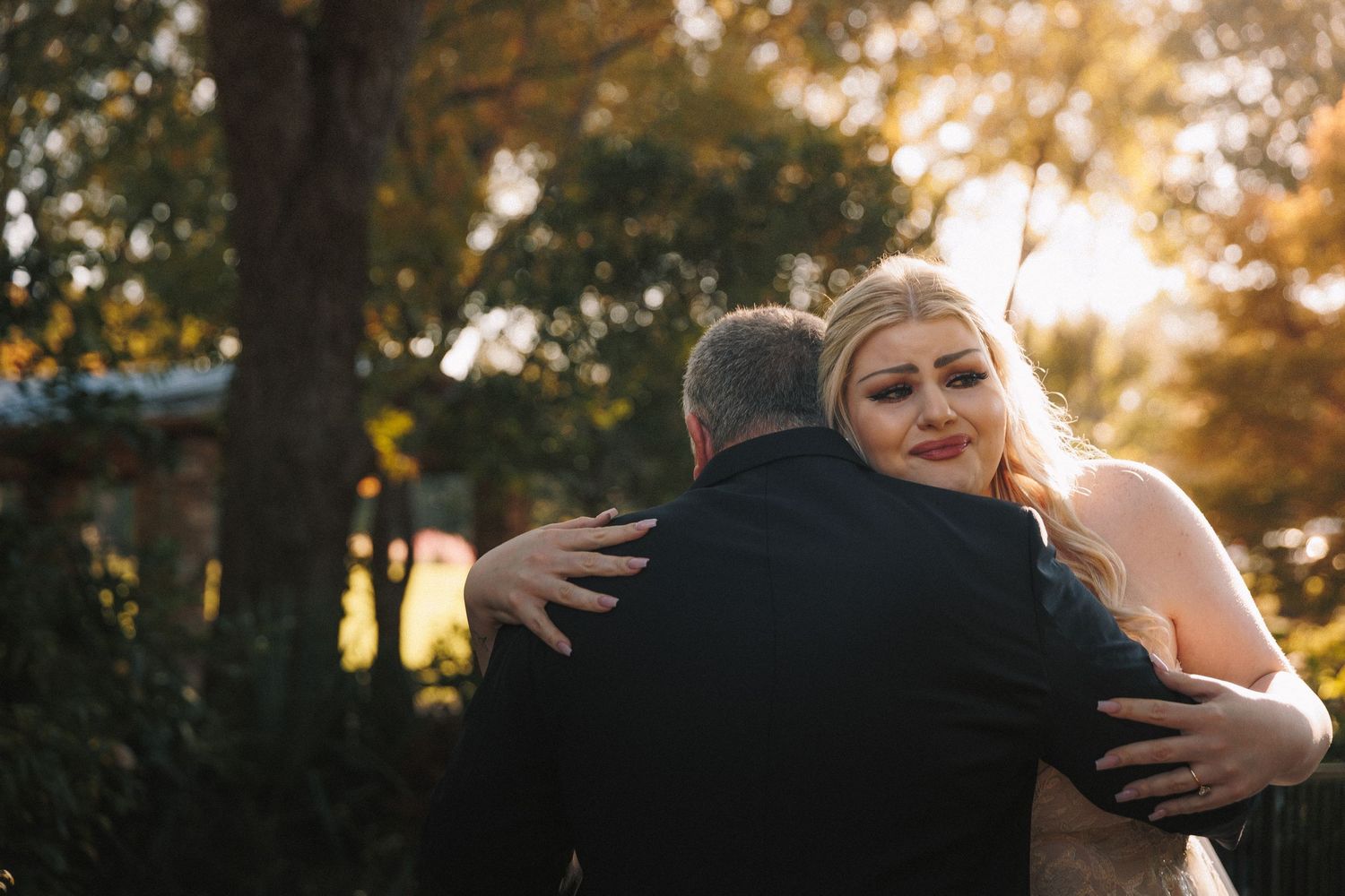 Emotional blonde bride in lace gown sharing a heartfelt hug with an older man outdoors at golden hour.