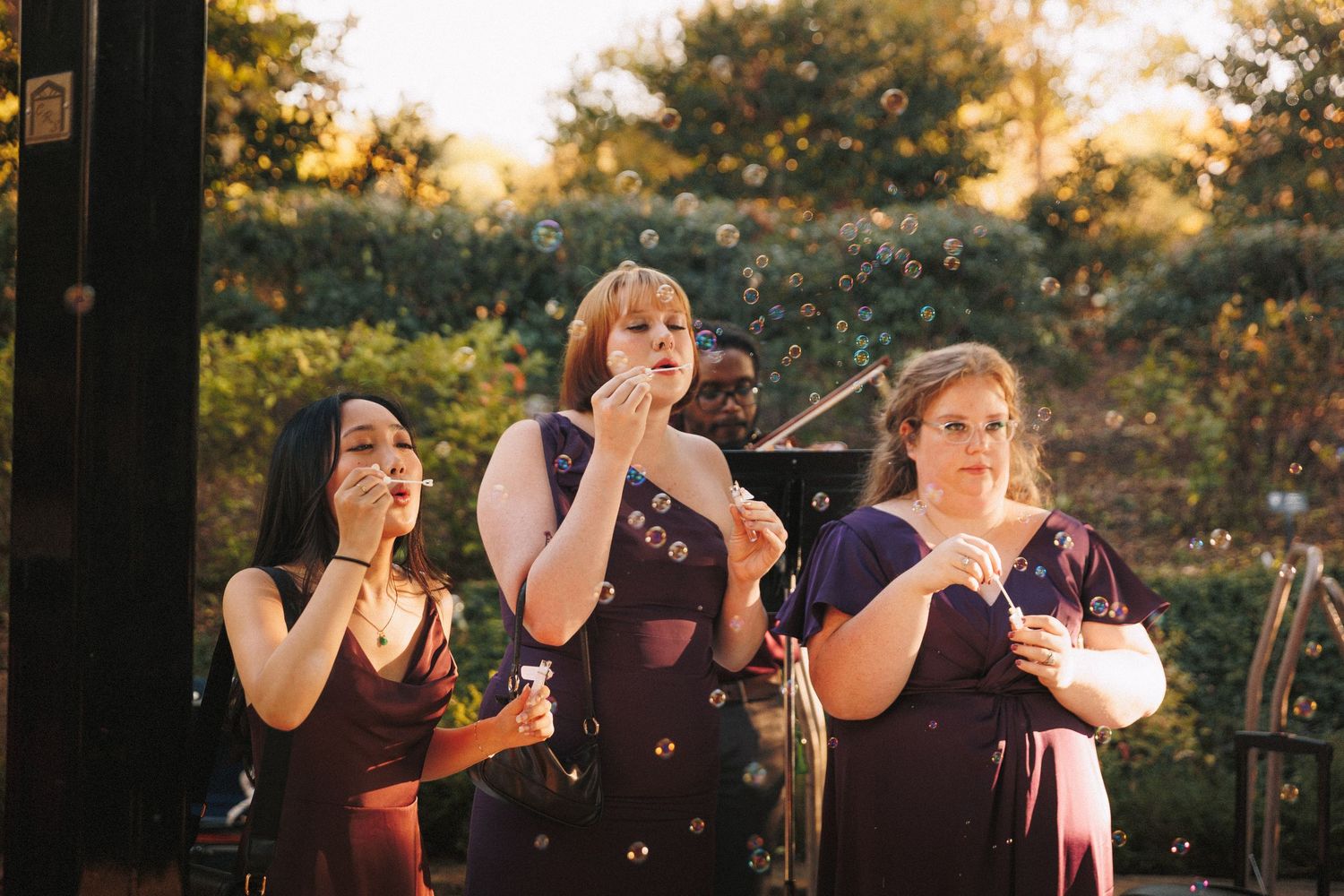 Three bridesmaids in burgundy dresses blow bubbles at outdoor wedding reception at golden hour.