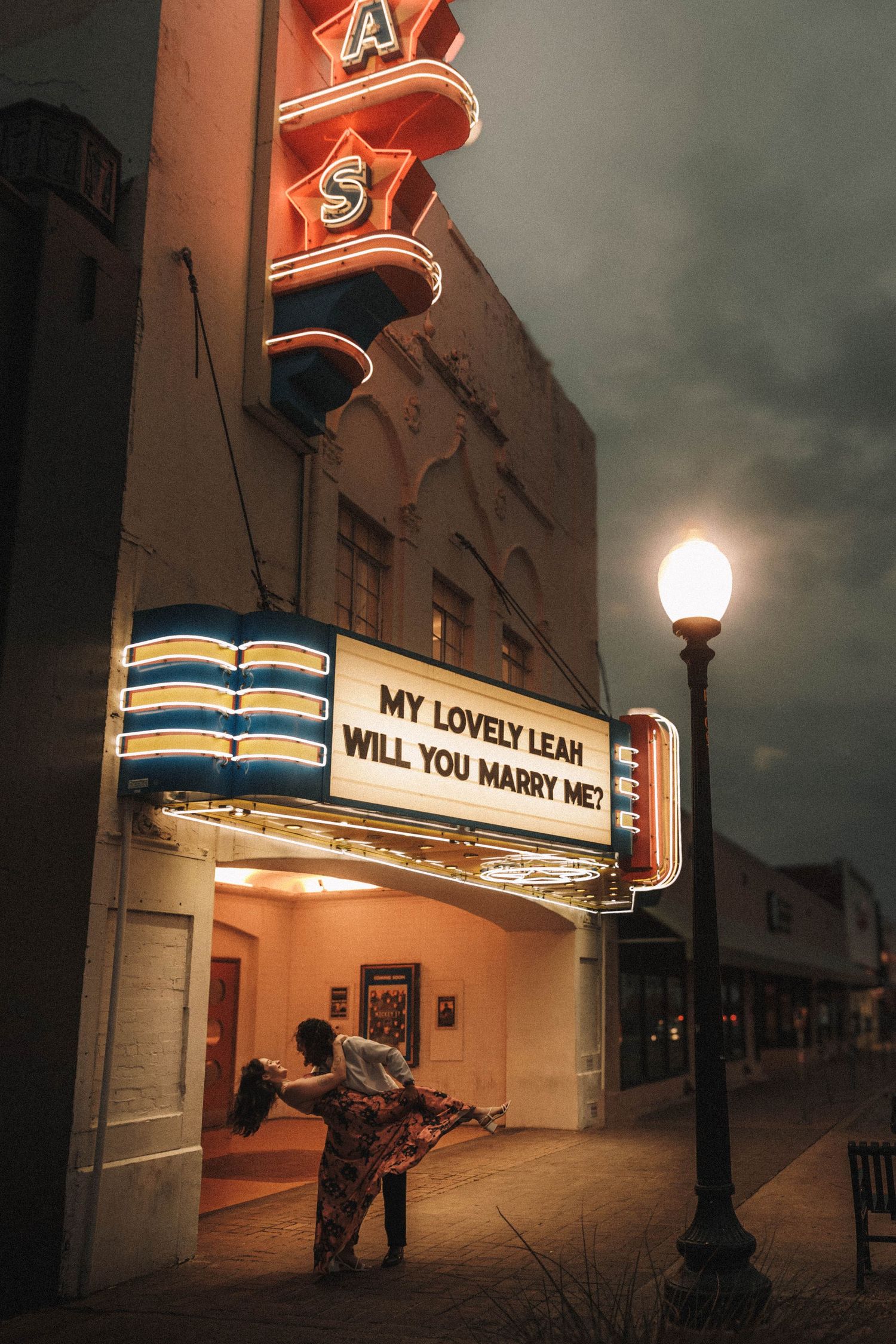 Couple embraces outside a classic theater marquee displaying a marriage proposal at dusk.
