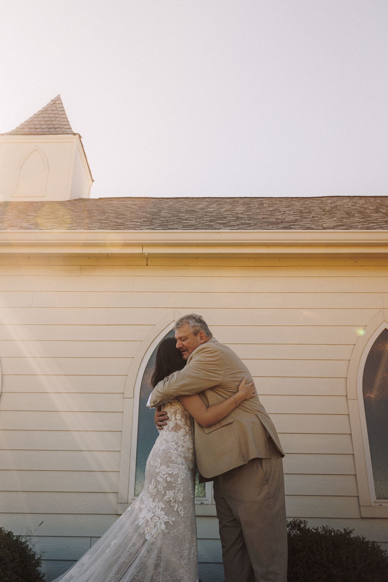 Bride and father share emotional hug outside white chapel on wedding day.