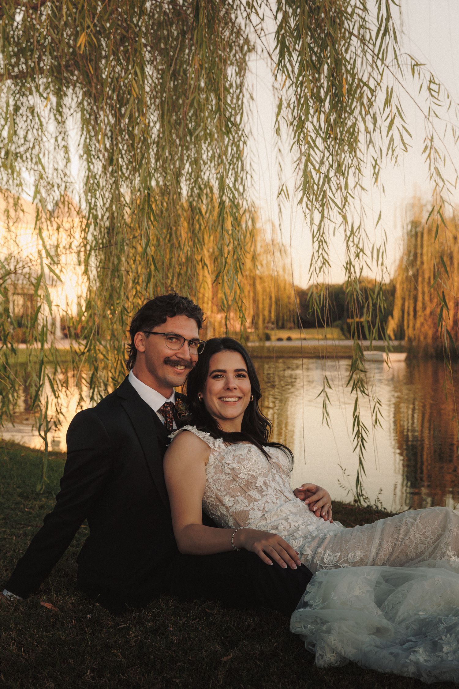 Smiling bride and groom sit together by pond under weeping willow at sunset.