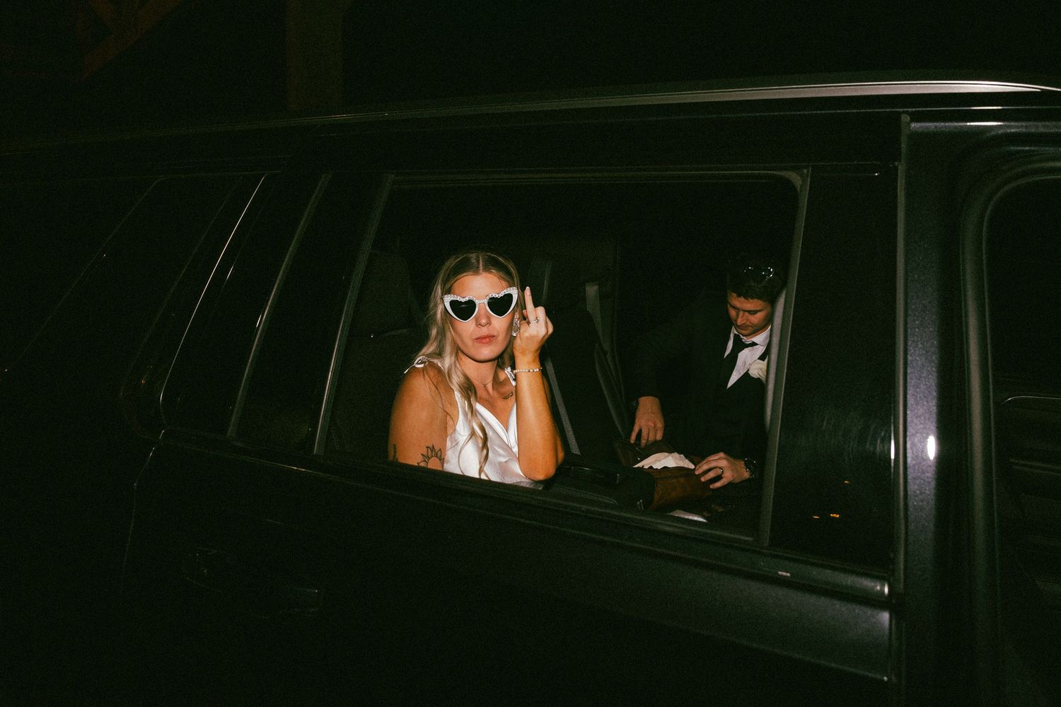 Smiling bride in sunglasses sitting in black car during wedding night getaway departure.