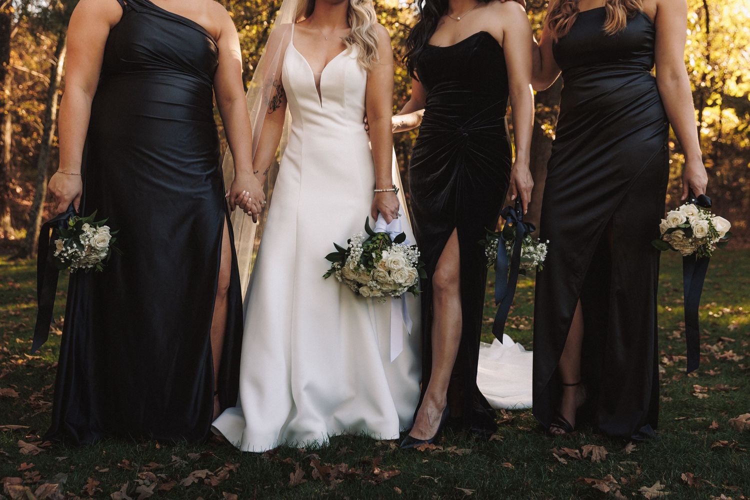 Bride in white gown with three bridesmaids in black dresses holding bouquets outdoors.