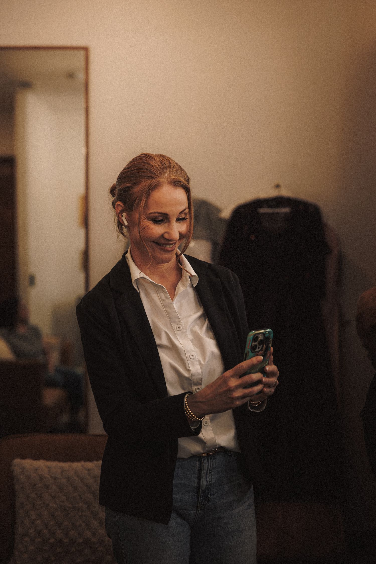 Smiling woman in black blazer and white shirt looking at her phone in a warmly lit room.