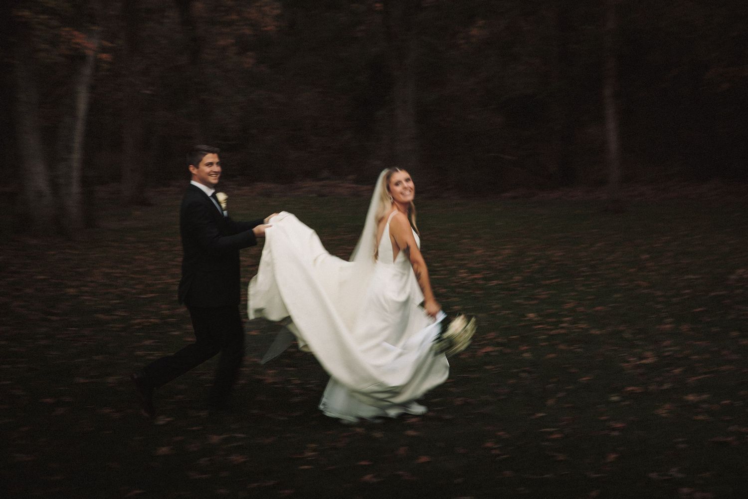 Bride in white gown twirling with groom in tuxedo in autumn forest setting.