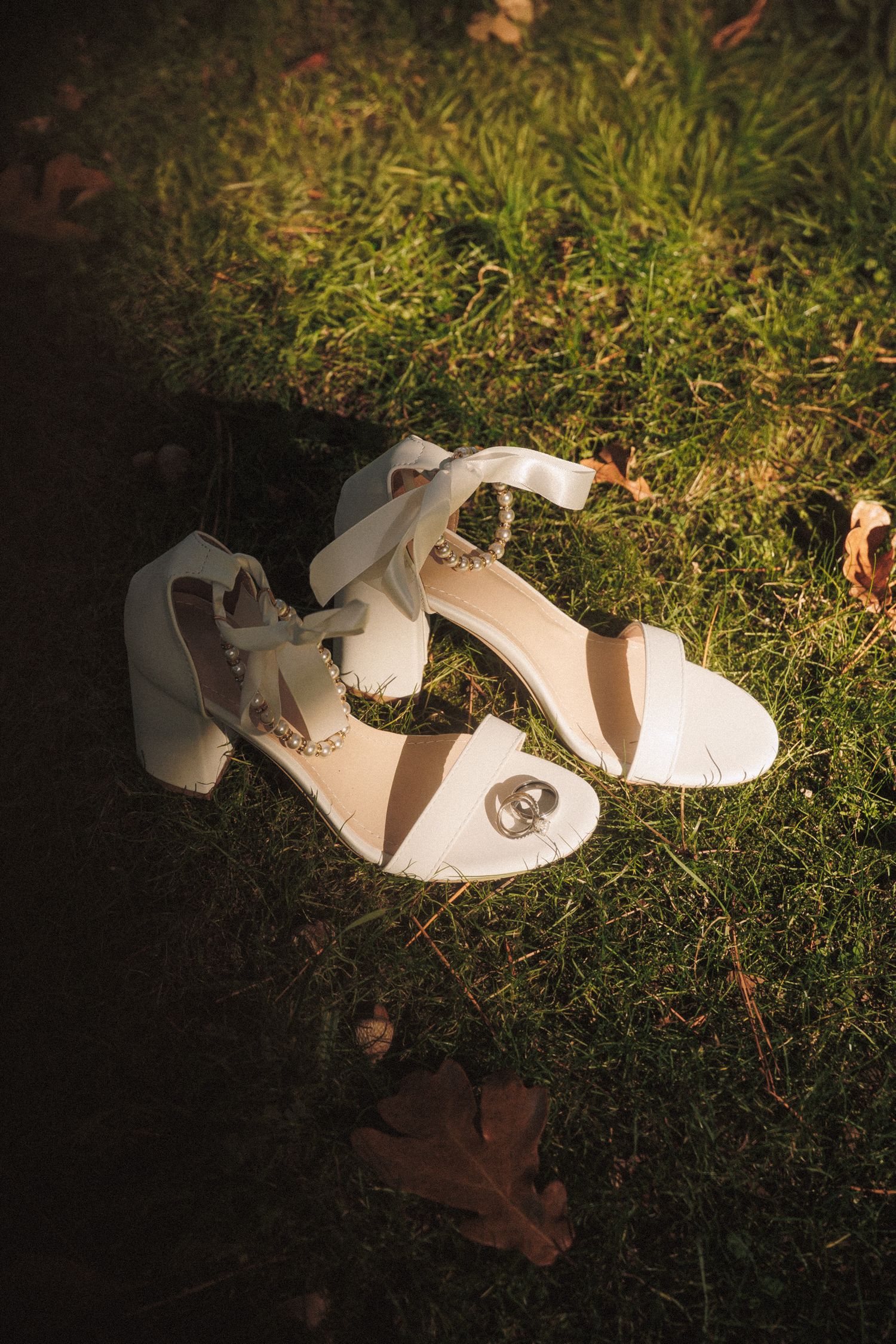White bridal heeled sandals with ankle ties resting on green grass with autumn leaves in warm sunlight.