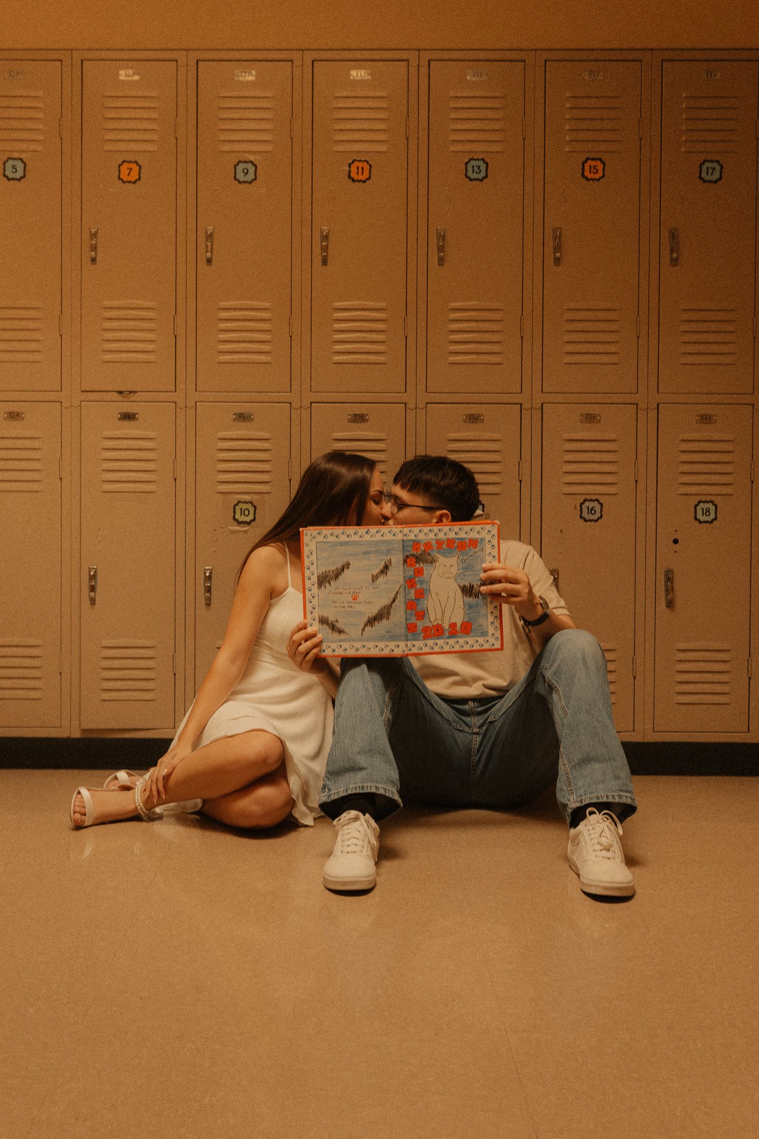 Couple sitting on floor in front of school lockers, sharing a kiss while holding a handmade card together.