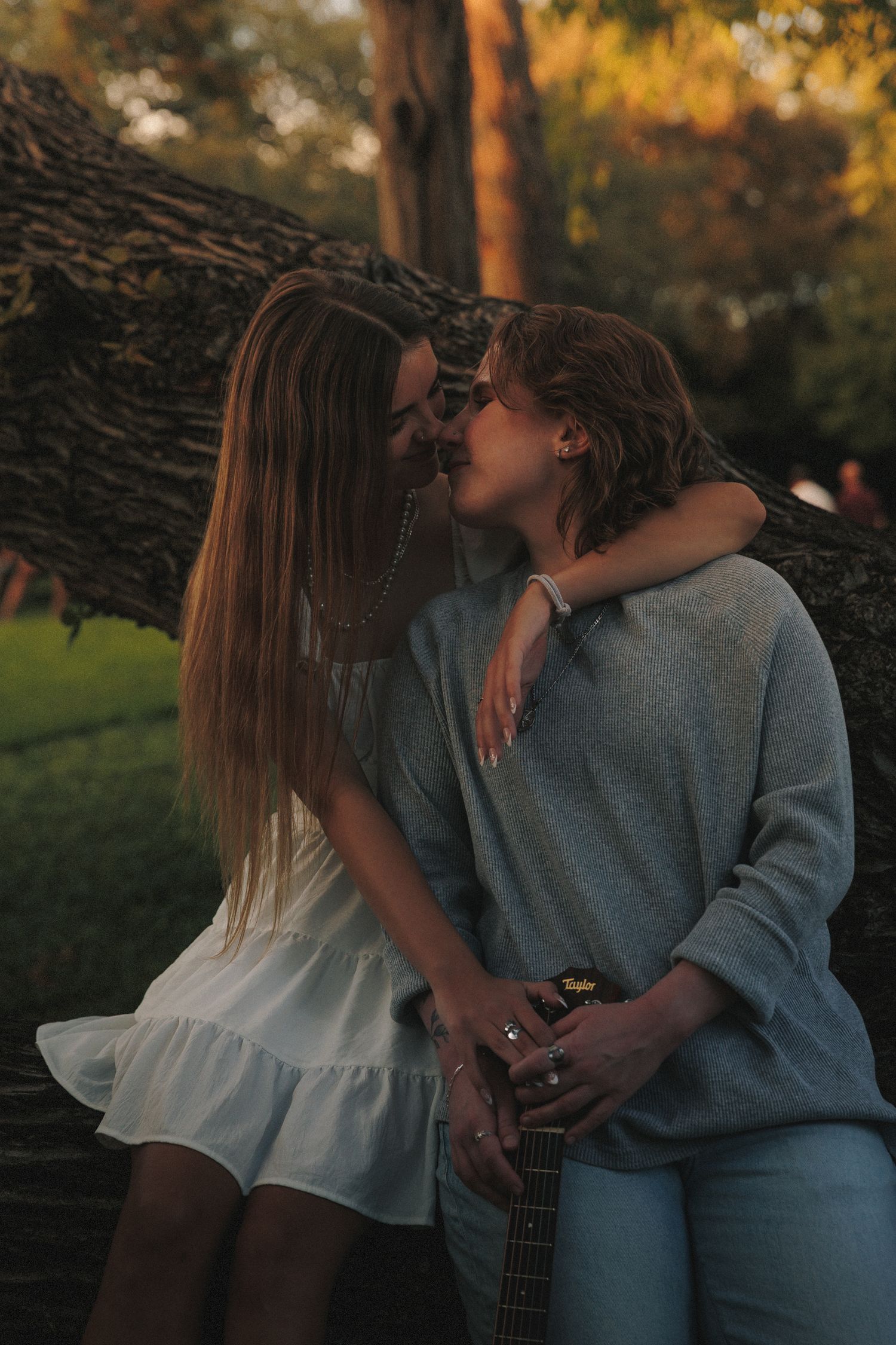 Two people in casual attire embrace near a lake during golden hour with a guitar beside them.