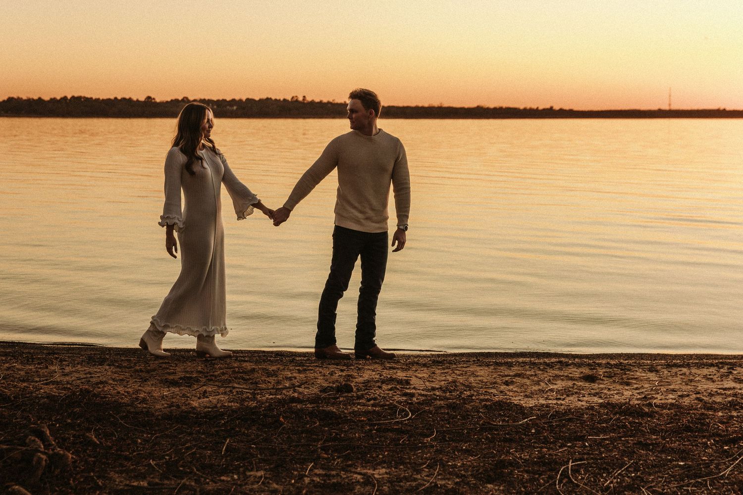 A couple walks along a tranquil beach shoreline at sunset creating silhouettes against the golden sky.